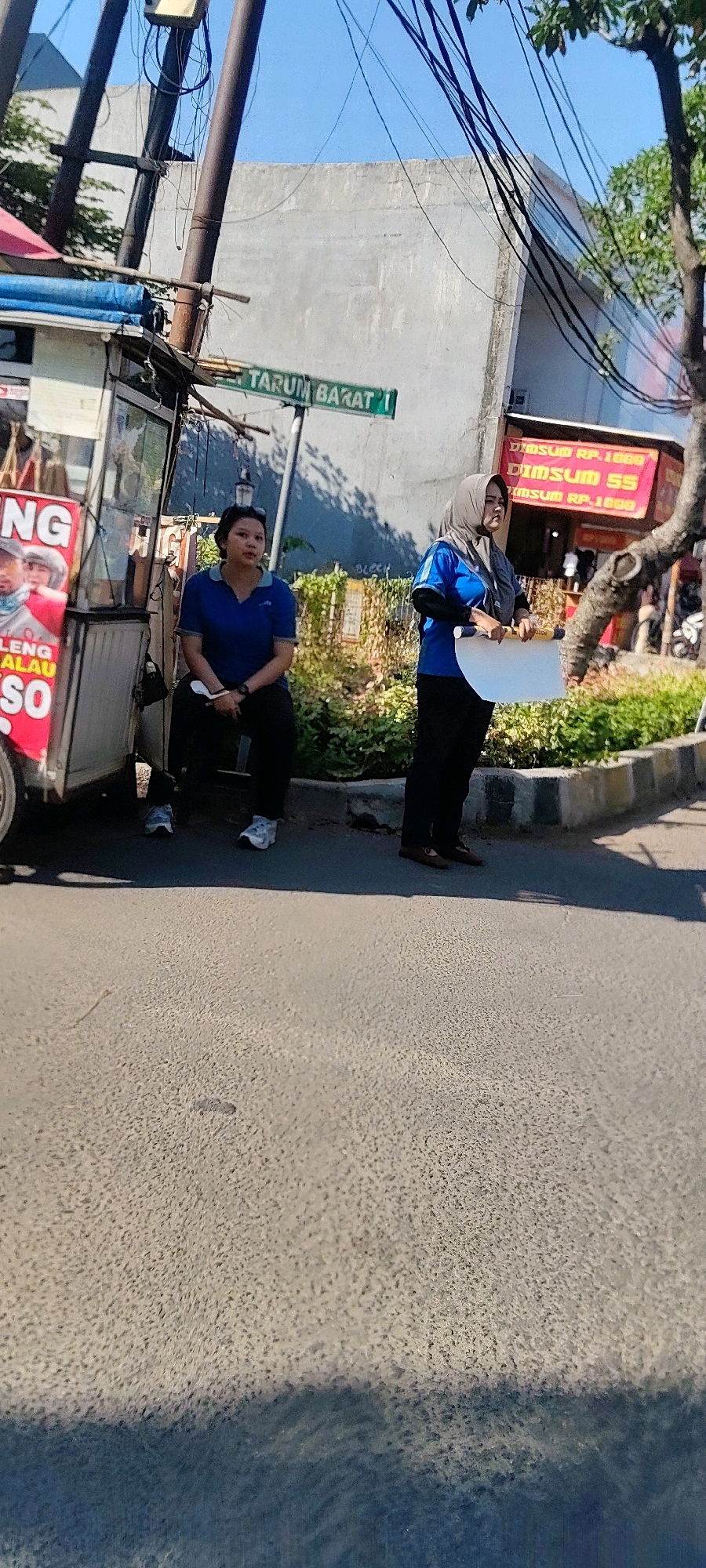 The image depicts a street scene in Cibatu, Indonesia, on a bright, sunny day. Two women are visible. The woman on the left, wearing a blue polo shirt and dark pants, is seated near a food cart. The food cart has a pink advertisement on its side. The woman on the right, wearing a blue shirt and a grey hijab, stands and holds a white piece of paper and a yellow object, possibly a banner or a marker. Behind them, a green street sign reads "TARUM BARAT 1". In the background, a building with a grey facade is visible, along with power lines. A tree with green foliage is also present. The ground is paved with asphalt. A building with a red sign advertising "DIMSUM" is also visible in the background.