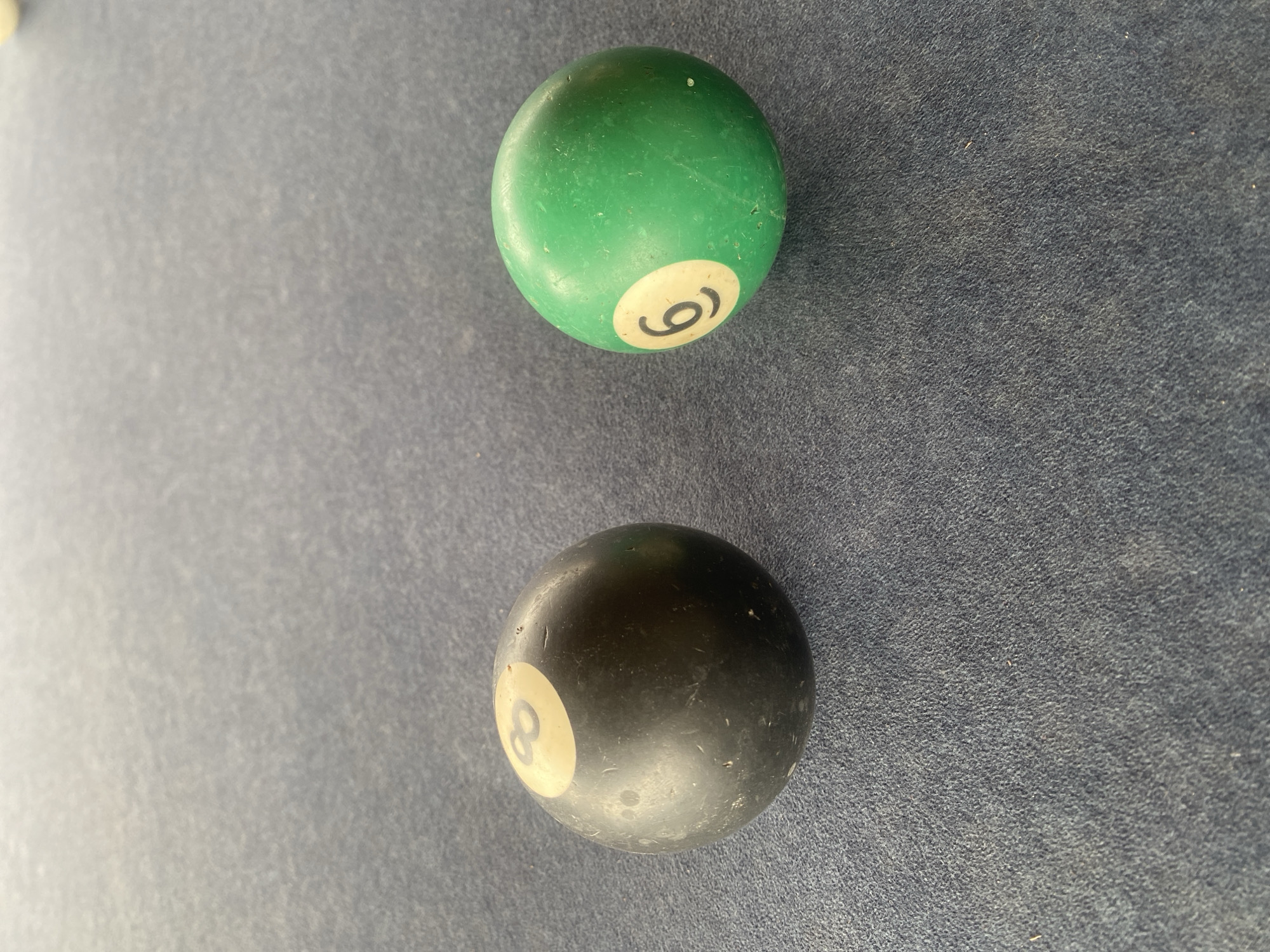 The image shows two pool balls on a blue felt surface, likely a pool table. The ball at the top is green with the number 6 visible. The ball at the bottom is black with the number 8 visible. The location in the city of Vom, Nigeria is unknown. No people are present. There are no visible actions or interactions.