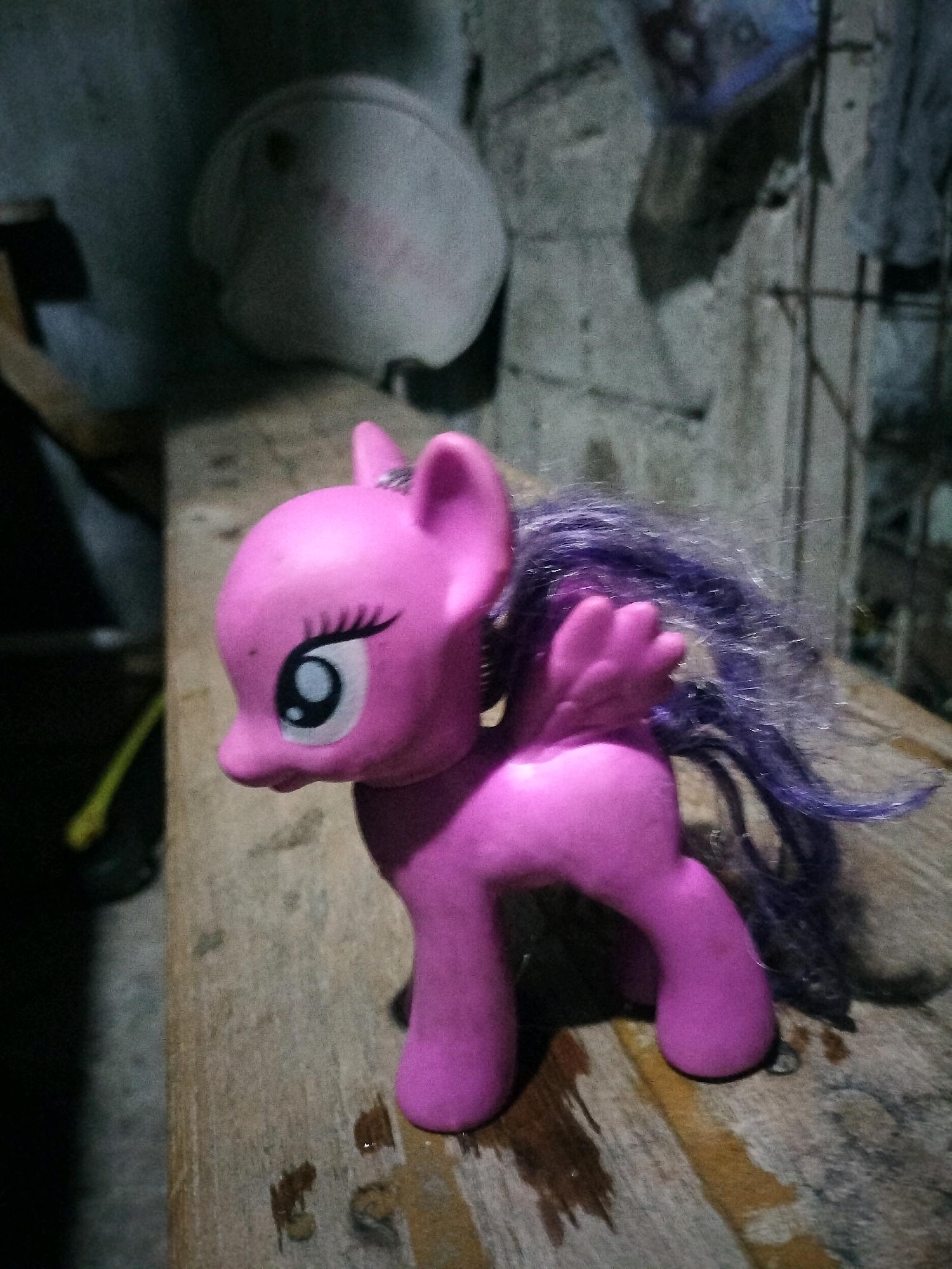 The image shows a pink toy pony with purple hair, placed on a wooden surface. The pony has large eyes with black eyelashes and white inner circles. Its body is facing towards the left, with its head turned slightly down. There are also some objects and a concrete wall in the background. The lighting is dim, suggesting an indoor or evening setting. The context of South Jakarta, Indonesia is not visually represented in the image.