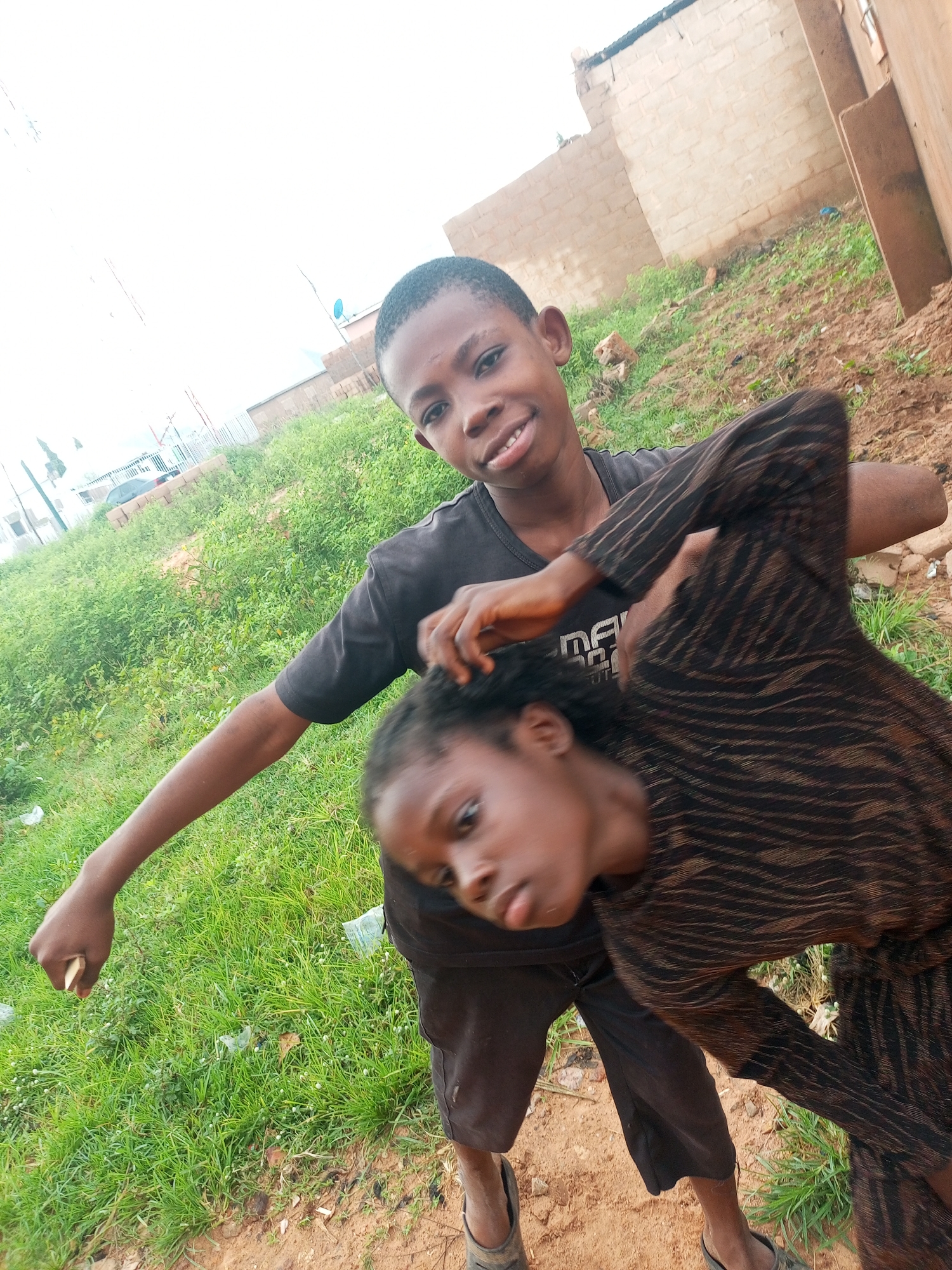 A boy, appearing to be around 10-12 years old, wearing a black t-shirt and dark shorts, is captured from a low angle. His left arm is extended, holding a small, light-colored object. He is looking towards the camera with a slight smile. A girl, younger than the boy, with dark hair and wearing a patterned dress, is positioned in front of him, her head tilted down and her face partially obscured, suggesting she is being playfully held or tickled by the boy. Both children are standing on dirt and grass, with some scattered debris. In the background, there are brick structures and what appears to be a fence or railing. The sky is overcast and bright, suggesting daylight. The scene takes place outdoors, in a somewhat undeveloped area, possibly within the city of Tempure, Nigeria, given the context of the prompt.