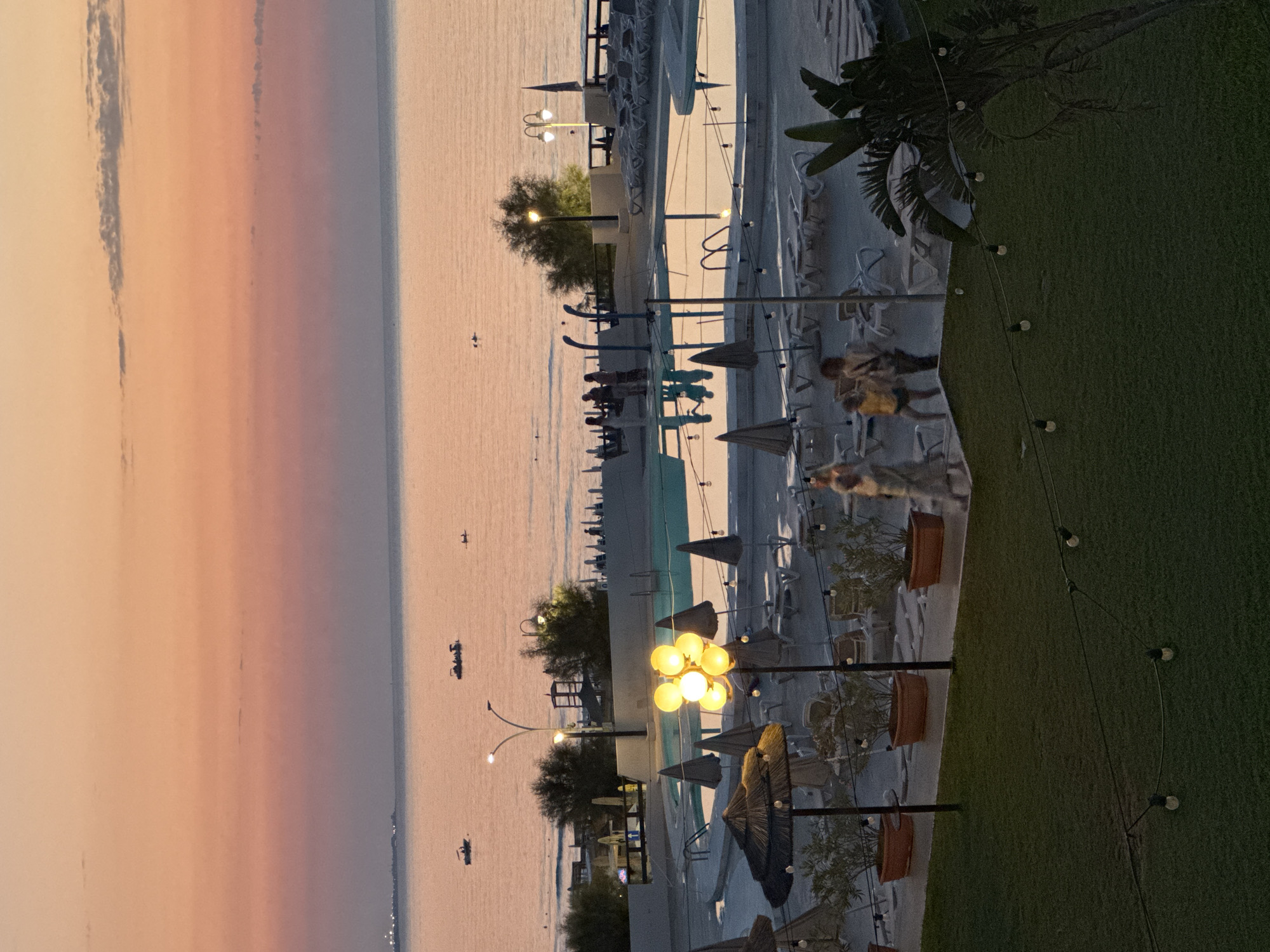 The image depicts a twilight scene at a resort or hotel complex on the coast of Isola delle Femmine, Italy. The sky is painted with soft hues of orange and pink, transitioning to a pale purple near the horizon. The sea is calm, reflecting the colors of the sky, with several small boats visible on its surface.
In the foreground, a swimming pool area is set up with lounge chairs and umbrellas. A group of people, appearing to be guests, are strolling around the pool area. Some are walking towards the pool, while others are walking away. There is a prominent cluster of bright yellow lights illuminating a section of the pool area. String lights are also visible, strung across the scene, adding to the ambiance. Adjacent to the pool, there is a well-maintained lawn. Further in the background, more lounge chairs are stacked, and some vegetation, including trees and potted plants, are present. The architecture of the resort is visible, characterized by white structures and railings. The overall impression is one of a peaceful evening at a seaside resort.