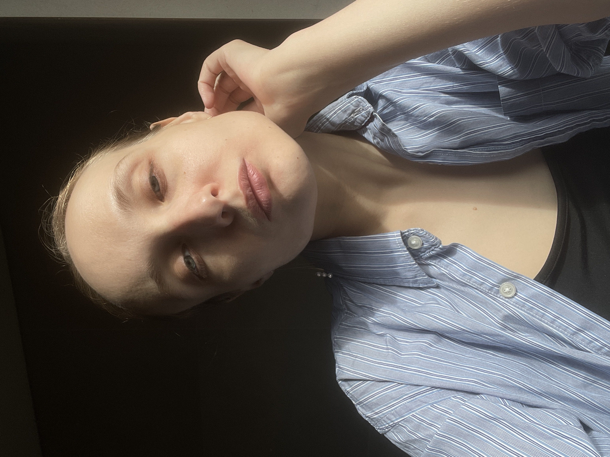 An indoor portrait features a young individual with fair skin and light hair, looking directly at the camera with a neutral to contemplative expression. The subject's head is tilted significantly, and a hand is gently placed near their right ear. They are wearing a light blue and white striped button-up shirt, partially unbuttoned, over a black undershirt or top. A delicate necklace with three small, bead-like elements is visible around their neck.
The lighting is high-contrast, with a strong light source from the upper right illuminating the subject's face, arm, and parts of the shirt, creating prominent highlights on the forehead, nose, and cheek, and casting deep shadows across the left side of the face and neck. The background is almost entirely dark and indistinct, offering no specific environmental details. The lips are slightly parted and appear to have a light pink or mauve tint. No text is visible in the image.
The image suggests a posed photograph, possibly a selfie. The location context provided is Подшивалово, Russia.