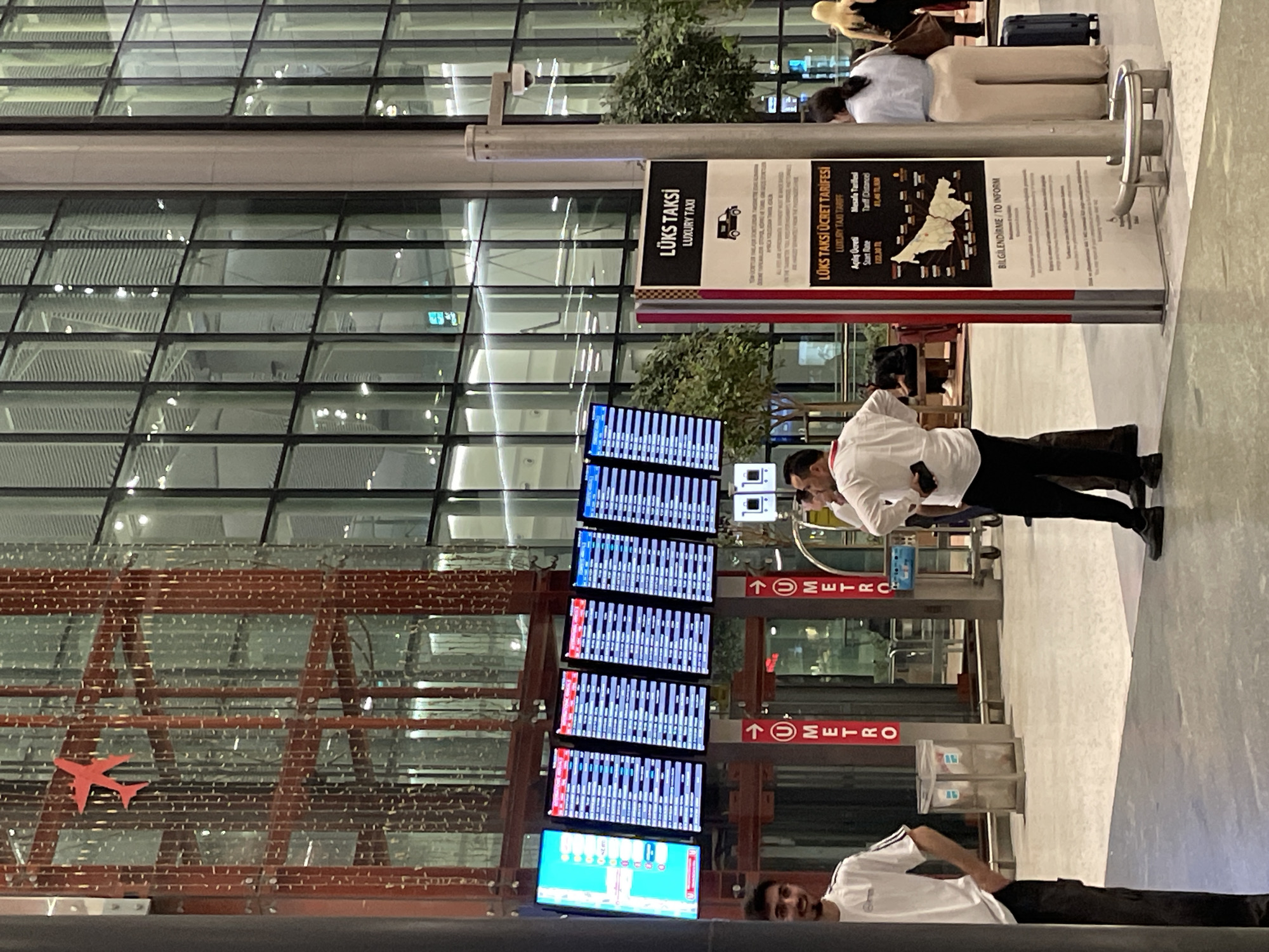 The image shows a modern airport interior with high ceilings and large glass windows. In the foreground, several digital flight information boards display departure and arrival times. To the right, a sign for "LÜKS TAKSI" (Luxury Taxi) is posted. People are seen standing and walking around the terminal. The overall environment suggests a busy transportation hub. The location is identified as Arnavutköy, Turkey, which is home to Istanbul Airport.