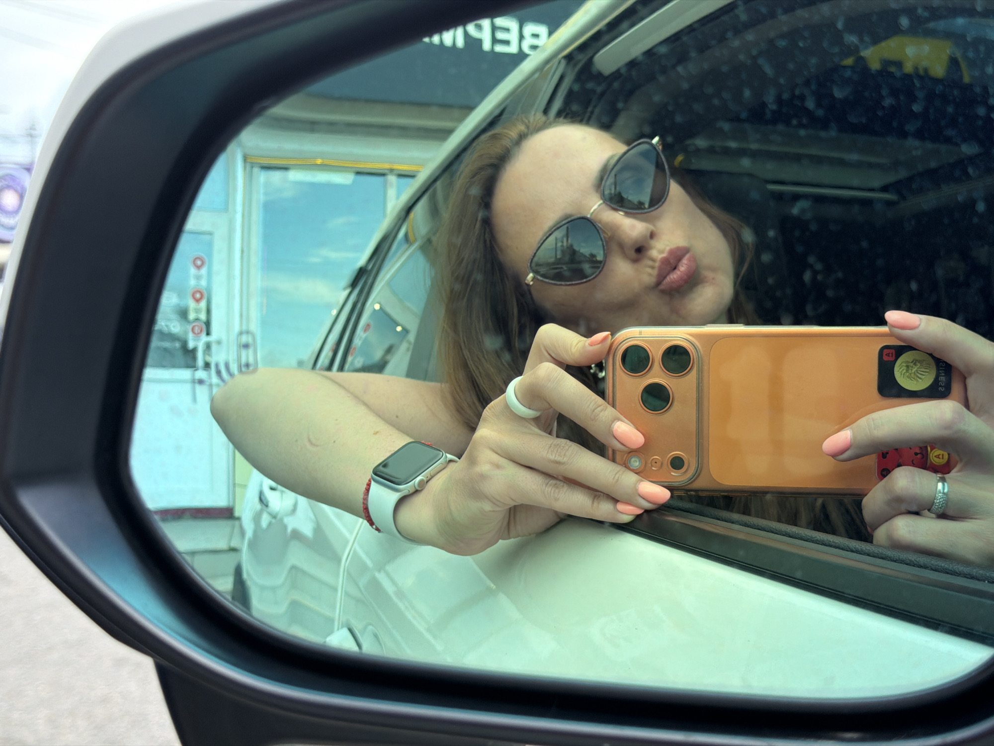 The media file is a close-up, reflected selfie taken by a woman inside a car. The woman is wearing sunglasses and has her lips puckered as if blowing a kiss. She is holding an orange smartphone with a multi-lens camera system in her right hand. The phone appears to have a sticker or case on the back with a design that includes a lion's head. She is also wearing a white ring on her left hand and a smartwatch with a white band and red string bracelet on her left wrist. Her fingernails are painted a peach color.
The background, as seen in the car's side mirror, shows a bright, possibly daytime scene. There is a building with glass windows and doors, and some signage is visible on the building. The reflection also shows a part of the car's interior, including the ceiling and some window condensation or dirt. The overall impression is that the photo was taken outdoors, possibly at a drive-through or a stop along a road. There are no explicit weather cues, but the lighting suggests it's not dark.
A notable detail is the text "BEP" visible in reverse in the reflection on the car window. Given the provided location context of Birobidzhan, Russia, this text might be part of a Russian word or phrase. The woman's facial expression and the selfie-taking action suggest a lighthearted or playful mood.
