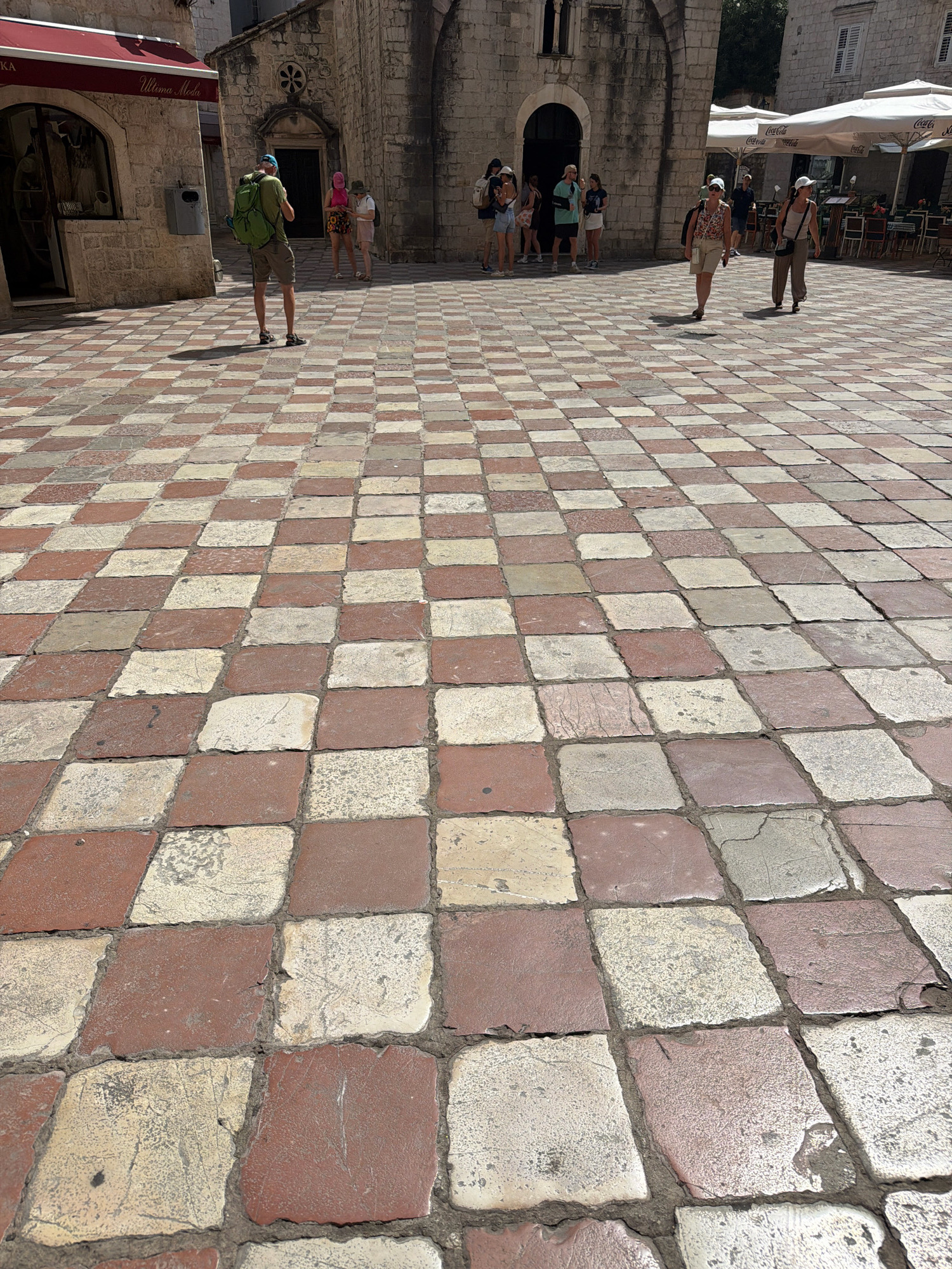 The image shows a view of a paved square in Kotor, Montenegro. The pavement is laid in a checkerboard pattern of reddish-brown and beige square tiles. In the background, there are stone buildings with arched entrances and windows. Several people are visible in the square. On the left, a man wearing a green backpack and shorts is standing facing away from the camera. Further back, a group of people, including women in light-colored clothing and hats, are gathered near a building entrance. To the right, two women are walking across the square, one in a floral top and shorts, the other in a beige top and trousers. In the far right background, outdoor seating with tables and umbrellas is set up, suggesting a cafe or restaurant. The overall scene depicts a sunny day in a historic European city square.