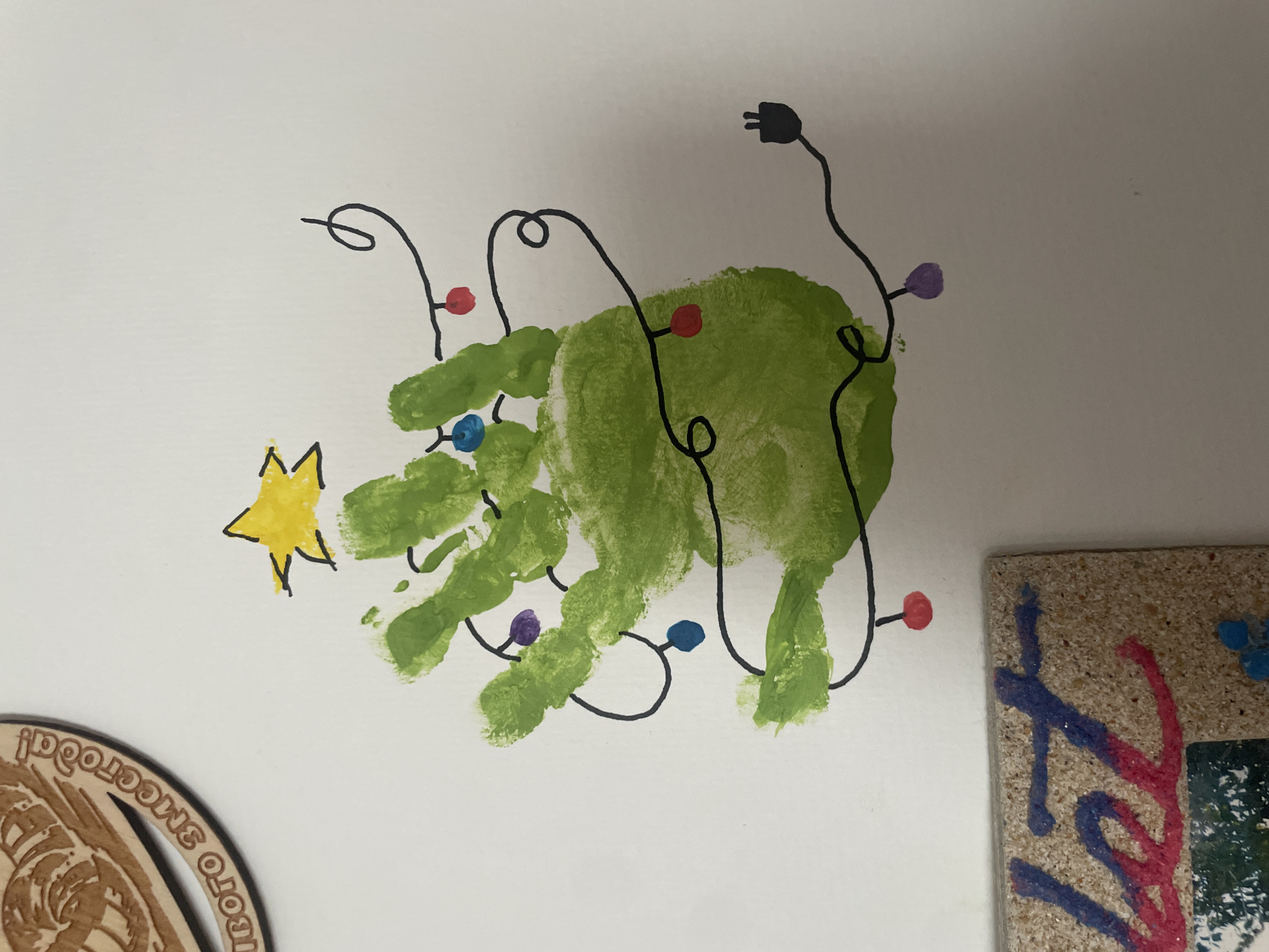 A child's artwork is displayed on a white wall. The artwork depicts a Christmas tree made from a green handprint, decorated with a string of lights with multi-colored bulbs (red, blue, purple). A yellow star is located to the left of the tree.
Parts of a wooden medallion with what appears to be Cyrillic text reading "...ОГОГО SMCCРОГОДA!" and the edge of a framed photo or artwork with colorful paint streaks are visible near the handprint Christmas tree. The location information is not present in the picture.