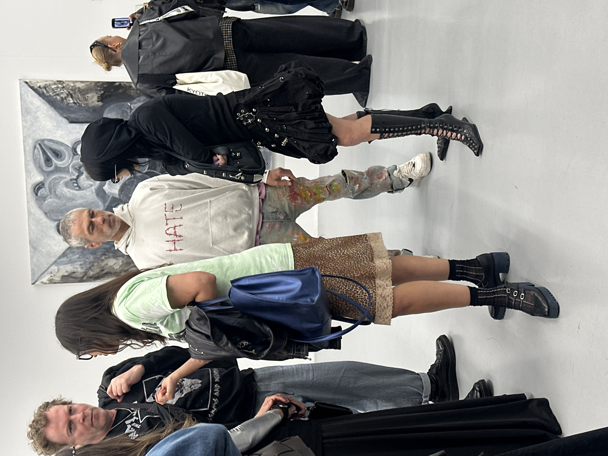 The image, correctly oriented, captures a group of people standing in what appears to be an art gallery or exhibition space in London, United Kingdom. The setting features clean white walls and a light-colored floor, creating a bright and minimalist environment.
A prominent feature of the scene is a large, black and white abstract or expressive painting displayed on the wall behind the central figures.
In the foreground, a person with curly light brown hair is visible, wearing a black graphic t-shirt and blue jeans. Next to them stands an individual with long dark hair, dressed in a light green tie-dye t-shirt, a distinctive leopard print mini-skirt with a lace trim, and dark buckled shoes over plaid socks. They are carrying both a blue shoulder bag and a black backpack.
Further into the scene, a man with short grey hair and a beard wears a white hoodie with the word "HATE" embroidered in red on the front, paired with paint-splattered jeans and white sneakers. Adjacent to him, a person with long dark hair is styled in a black hooded top or jacket, a black ruffled skirt embellished with silver studs and buckles, and striking black thigh-high laced boots. This person carries a black handbag.
In the background, a person with blonde hair in a bun, wearing a black jacket over a plaid garment, is holding a smartphone, seemingly taking a photo or video. A white bag bearing the text "KYOTO" is also visible in this area, alongside another partially obscured figure.
The individuals appear to be casually standing, observing the artwork, or socializing within the exhibition space. The overall mood is contemplative and relaxed, indicative of an art event.