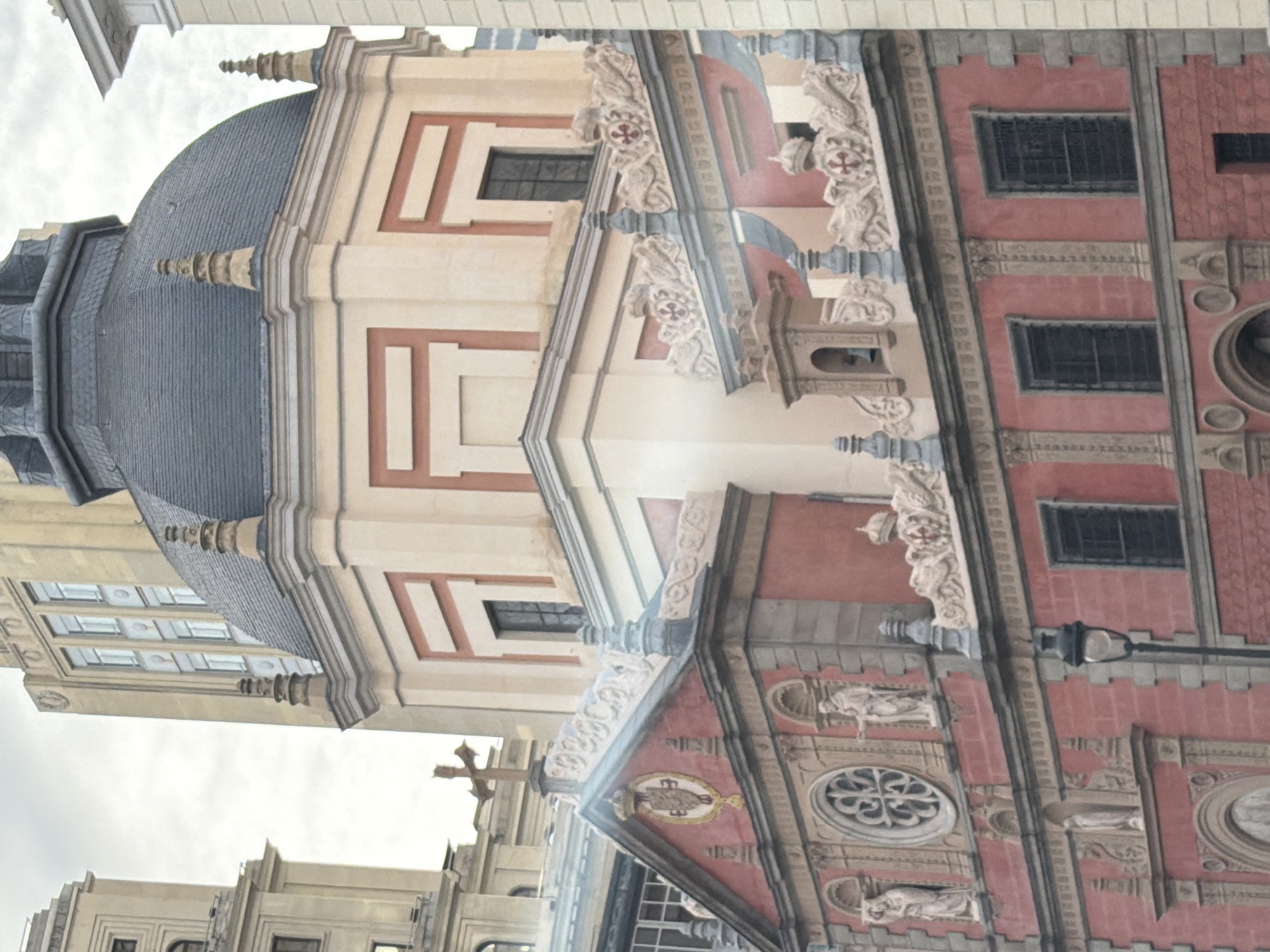 The image shows the facade of the church Iglesia de San Isidro, located in Madrid, Spain. The structure features a prominent dome with dark grey tiles, resting upon a cream-colored section of the building decorated with red and white geometric patterns. Adjacent to this is a red brick section with windows and decorative carvings of statues. A cross is visible on the building's apex.