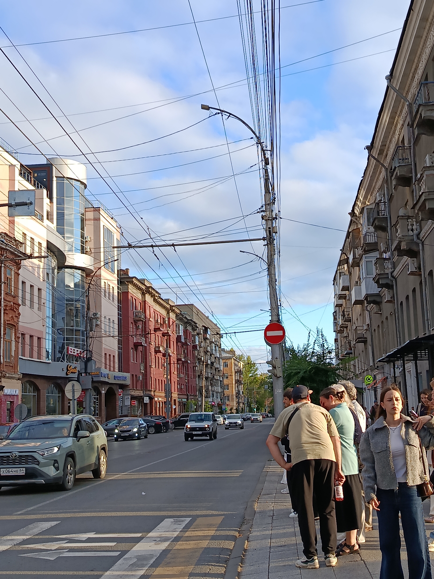The image depicts a street scene in Saratov, Russia, on a sunny day with a cloudy sky. In the foreground, a pedestrian crossing with arrows is visible on the asphalt. Several cars are driving on the street, including a light green SUV with a Russian license plate reading "X064HE 64". A group of people are standing on the sidewalk to the right of the street. On the left side of the street, buildings with multi-colored facades, balconies, and awnings line the road. Overhead, a complex network of utility wires crisscrosses the sky, supported by poles. One pole has a red circular "no entry" sign. The overall impression is a typical urban environment with infrastructure, traffic, and pedestrians.