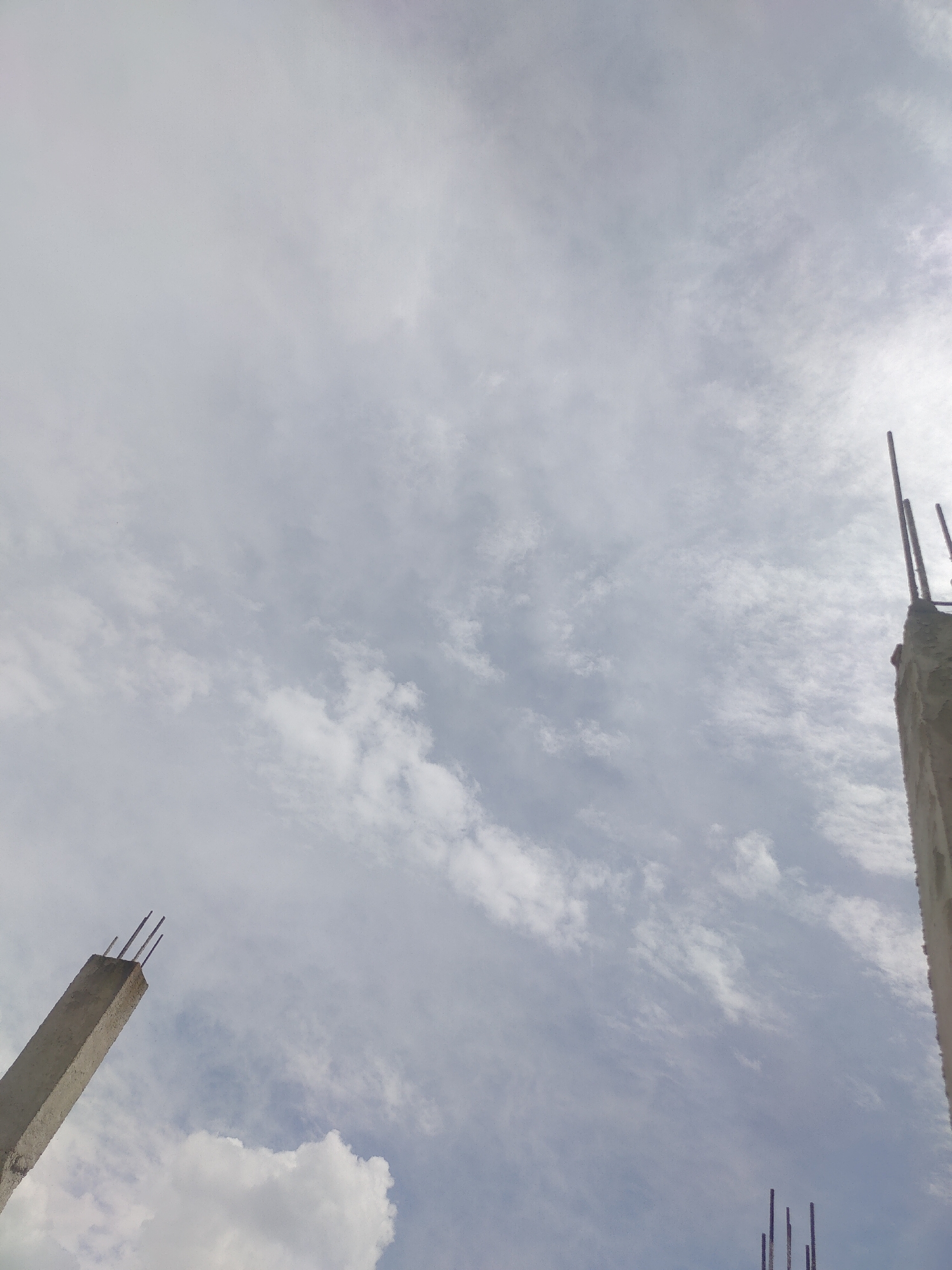 The image shows a cloudy sky with two concrete pillars, suggesting a construction site. The pillar on the left is leaning towards the left and has rebar protruding from its top. The pillar on the right is taller and more upright, also with rebar visible at the top. The clouds are thin and wispy, indicating partly cloudy conditions. The scene is likely an outdoor setting, possibly in a developing urban area. There is no specific indication of the city of Manali, India.