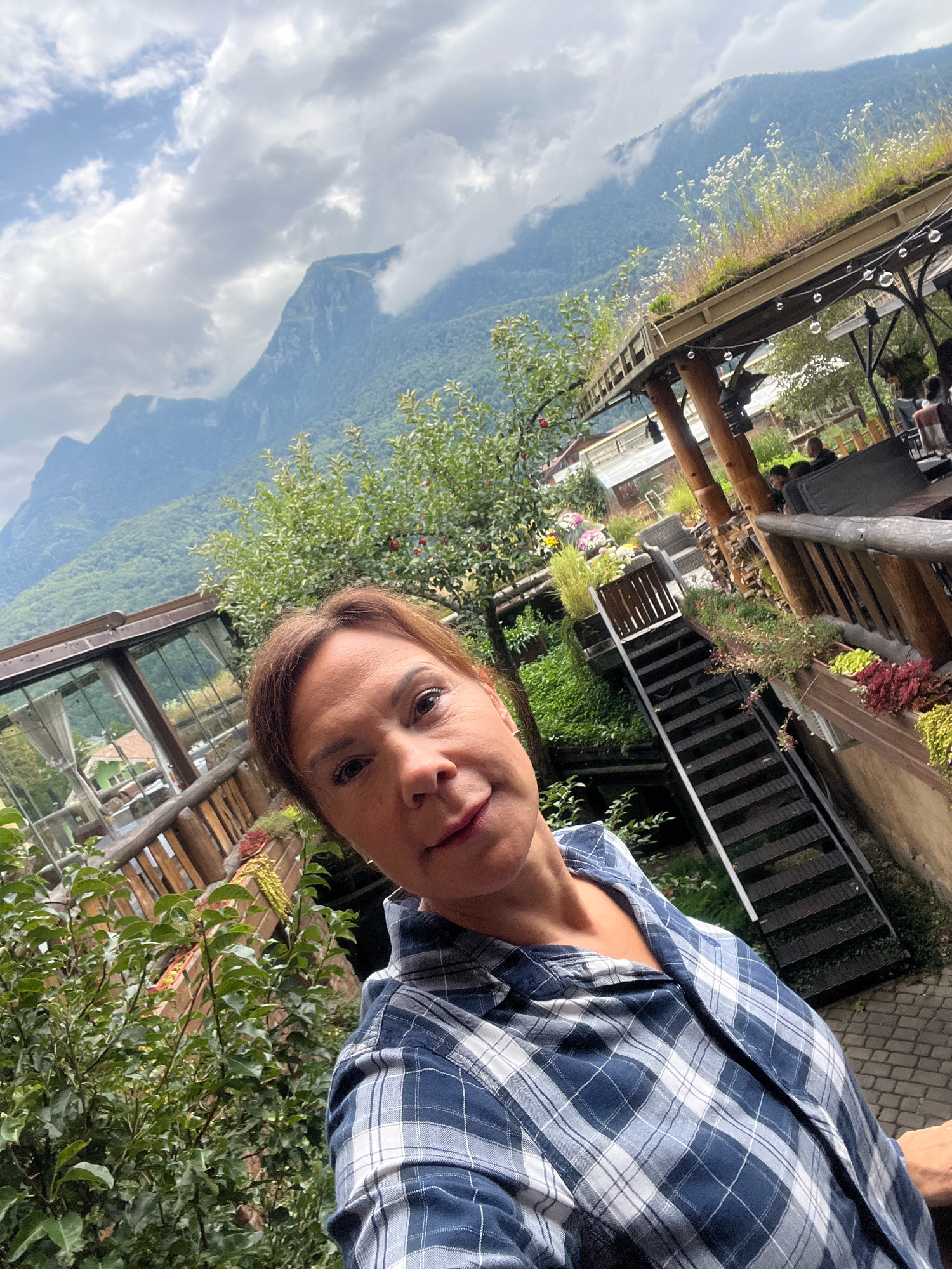 A woman with brown hair and fair skin is taking a selfie in an outdoor setting. She is wearing a blue and white plaid shirt. In the background, there are mountains, lush green trees, a building with a glass-covered section, and a wooden staircase leading to an elevated wooden structure with a green roof. Several people are visible in the background, seated at tables under the wooden structure. The sky is partly cloudy. The location is identified as Krasnaya Polyana, Russia.
