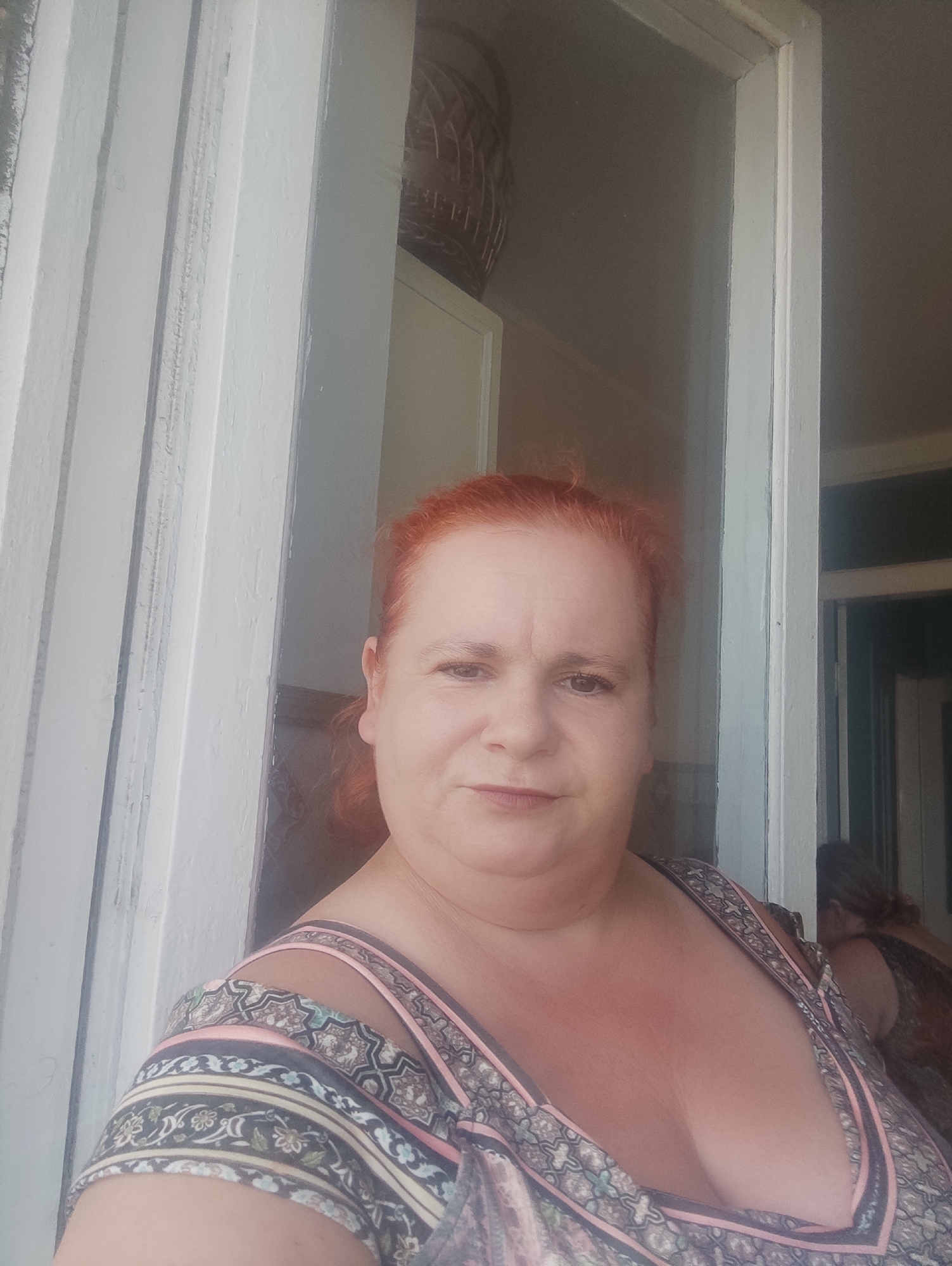A woman with red hair is taking a selfie indoors, in what appears to be a home with white wooden trim framing the scene. She is wearing a patterned dress with thin straps. In the background, another person can be seen in shadow. The lighting suggests it is daytime. The image does not provide any specific visual cues to identify the location as Chernivtsi, Ukraine.