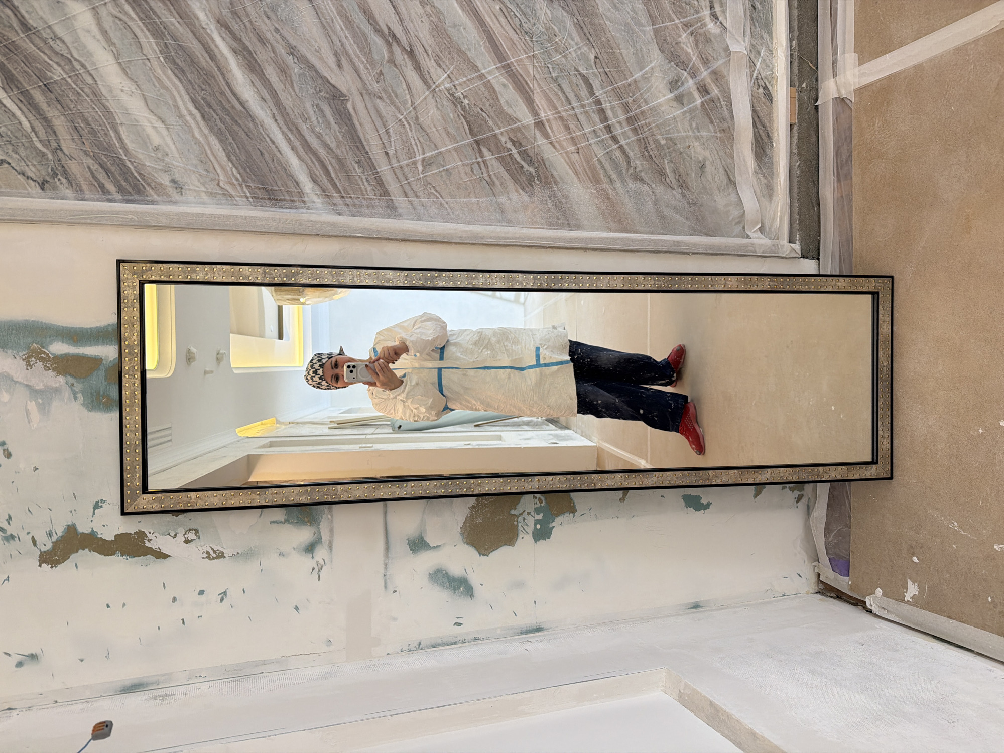 A person in a white protective suit and dark jeans is reflected in a rectangular mirror with a studded frame. The person is holding a smartphone and appears to be taking a selfie. The surrounding environment shows unfinished interior work, with walls partially painted and covered with plastic sheeting. The location appears to be a building under renovation, potentially in Khimki, Russia.