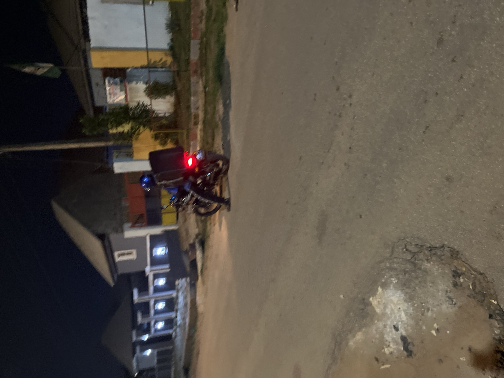 The image depicts a night scene on a paved road in Abuja, Nigeria. A person, wearing a blue helmet, is riding a motorcycle. The motorcycle is equipped with a black storage box, commonly used for deliveries. The rider's legs are visible, clad in dark pants. The motorcycle's rear brake light is illuminated red. In the background, there are buildings, one of which features a prominent Nigerian flag flying from a pole. The area appears to be a residential or commercial district, with streetlights and architectural elements visible. The road surface shows some wear and tear, with patches of debris and cracks.