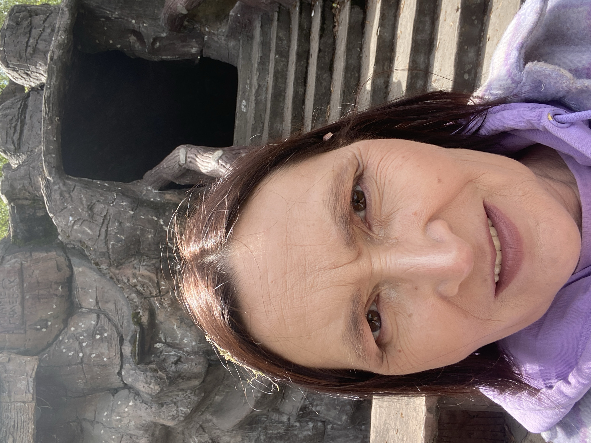 A close-up image shows a woman wearing a purple hoodie, positioned slightly to the side and rotated counter-clockwise. In the background, there's a dark cave-like opening constructed with gray stone, along with a set of concrete steps ascending. The location is identified as Novokuznetsk, Russia.