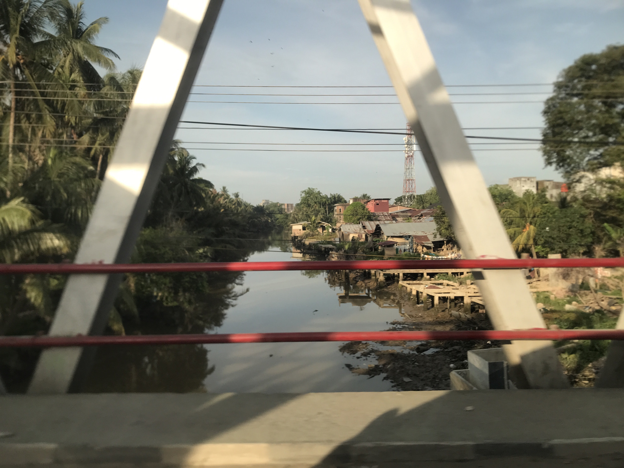 The image captures a view from a bridge overlooking a calm river or canal in Kotapantonlabu, Indonesia. Two prominent white diagonal girders of the bridge frame the scene, with red horizontal railings crossing the lower half of the frame. Below the railings, a concrete barrier is visible in the immediate foreground.
The narrow waterway, reflecting the sky and surrounding structures, dominates the midground. Its banks are lined with informal settlements and lush tropical vegetation. On the left side of the river, dense palm trees and other greenery create a thick canopy. On the right bank, a cluster of houses with varying roof types, some appearing to be built on stilts or elevated foundations extending over the water's edge, can be seen. The banks here also show exposed muddy areas and some debris.
In the background, on the right side, a tall red and white cellular tower stands out against the sky, surrounded by more trees and distant buildings. The sky is bright blue with a few scattered, faint clouds, and several small dark specks, possibly birds, are visible. The lighting suggests it is daytime, likely morning or late afternoon, with a soft, clear light illuminating the landscape. No people or visible text are present in the image.