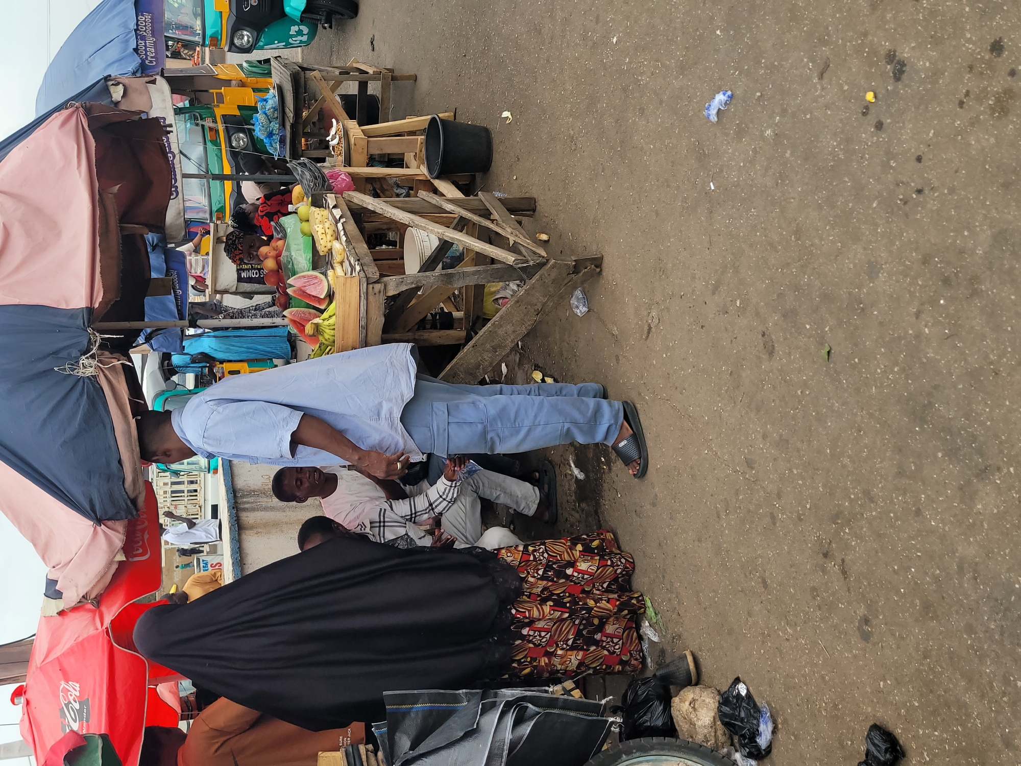 The image captures a street scene in Jos, Nigeria. A man in a light blue shirt and dark sandals stands near a makeshift fruit stand constructed from wooden planks, displaying watermelons, bananas, and other produce. Nearby, several people are seated. One woman is dressed in a black hijab and a colorful patterned skirt. There are Coca-Cola umbrellas and makeshift tent structures in the vicinity. The ground is paved but worn. In the background, there are yellow three-wheeled vehicles (keke napeps). Debris and litter are visible on the ground.
