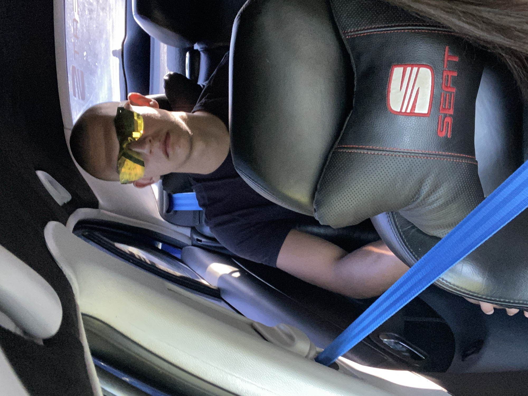 In Sevlievo, Bulgaria, a young man with a shaved head is pictured from the chest up, seated in the passenger seat of a car. He is wearing a black t-shirt and large, yellow, reflective sunglasses that obscure his eyes. The interior of the car is visible, with a black headrest for the seat in front of him, featuring red stitching and a white SEAT logo. A bright blue seatbelt is visible across his torso. The lighting suggests it is daytime, possibly with sunlight filtering into the car.
