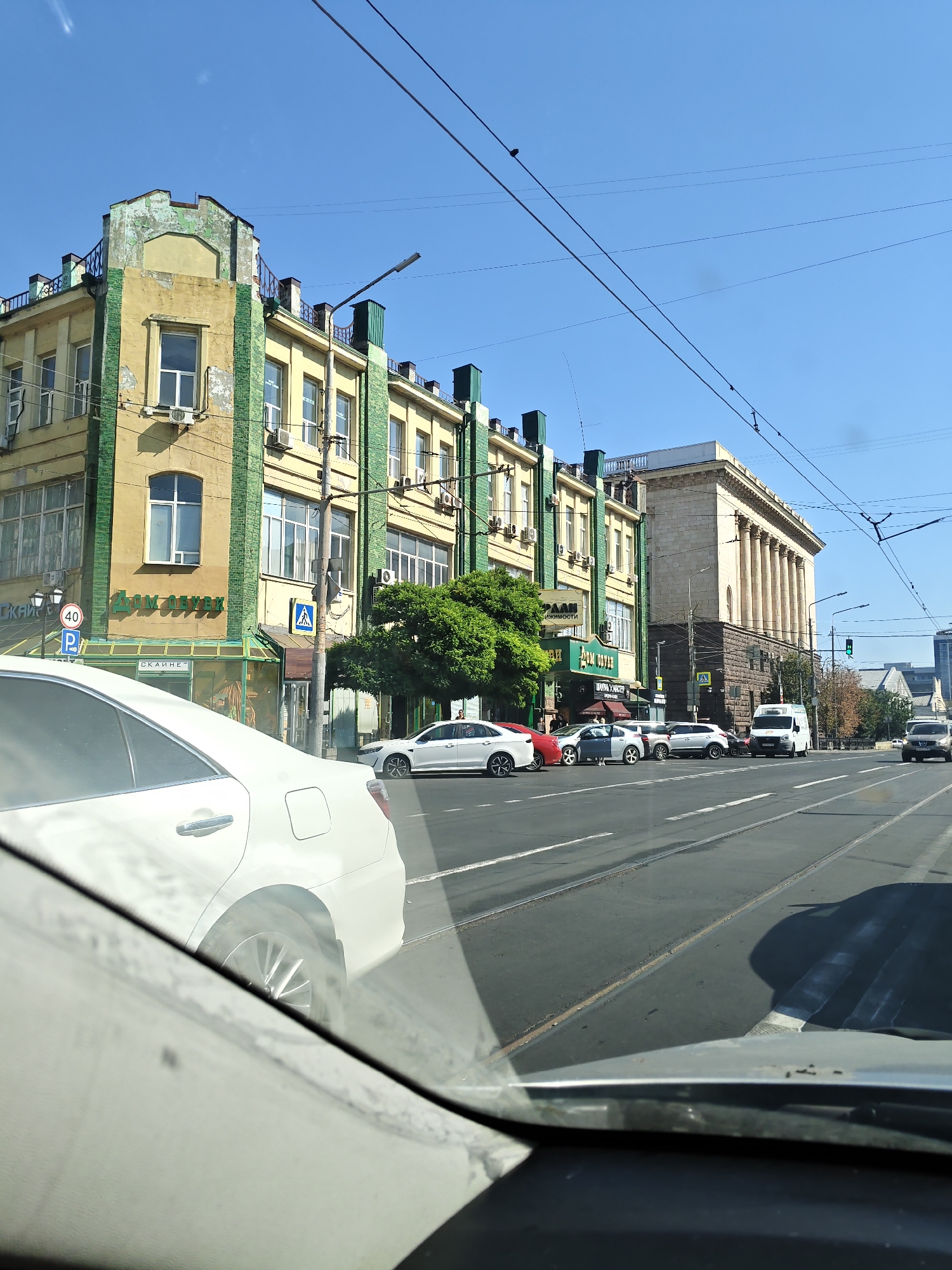 The image captures a view from inside a vehicle in Rostov-on-Don, Russia, looking out at a city street on a clear, sunny day. The street is a multi-lane road with tram tracks visible in the asphalt.
In the foreground, the white interior of the vehicle, including the windshield and dashboard, is prominent. A white car is partially visible on the left side of the frame, reflecting the sunlight.
Further down the street, several buildings line the right side. The building closest to the foreground has a yellow facade with green accents and architectural details, including several windows. A sign with the number "40" and a "P" for parking is visible on the left side of the street. Another building, appearing to be a commercial establishment with large glass windows, is situated next to it. Further down the street, a larger, classical-style building with columns stands out. A white van and other cars are driving or parked on the street.
A lush green tree is positioned between the commercial buildings. Electrical wires stretch across the blue sky, typical of an urban environment. The overall scene depicts a typical street view in a Russian city.