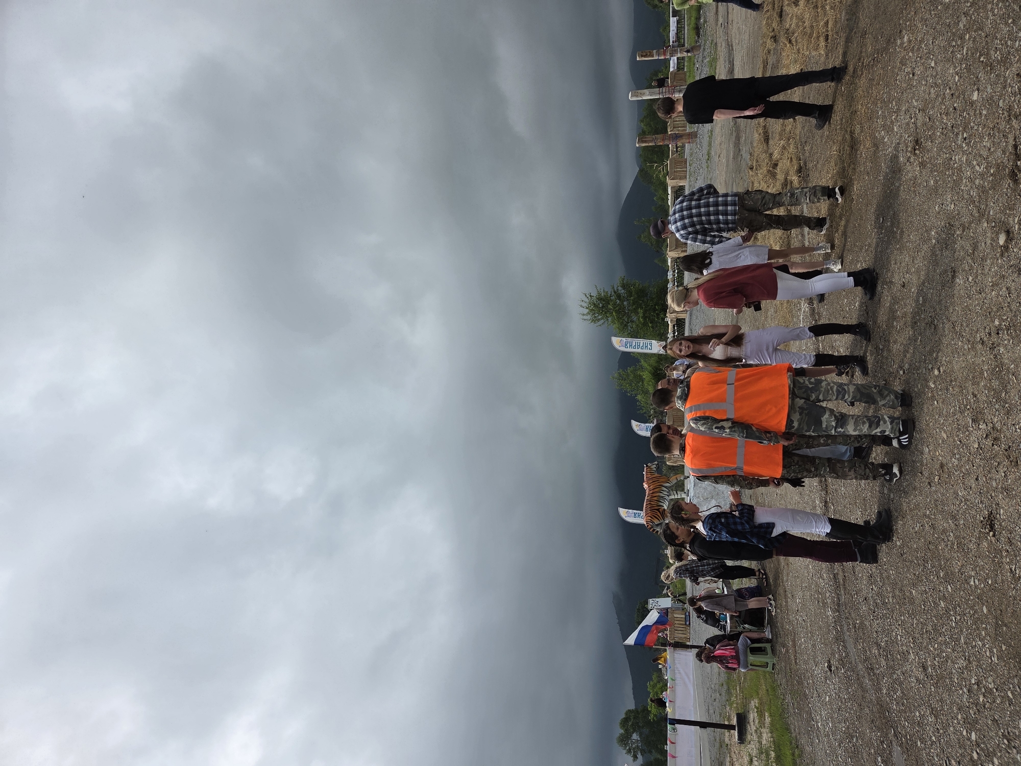 In Теплоозёрск, Russia, under a heavy, overcast sky, a group of people are gathered in an outdoor setting. The foreground is dominated by a dirt and gravel ground, with some scattered dry grass. Several individuals are standing, facing in various directions, some in conversation. Two men in the center foreground are wearing bright orange safety vests over camouflage-patterned clothing. To their left, a woman in a white top and white riding pants with tall black boots is facing away from the camera. To the right of the men in orange vests, a woman in a red top and white pants is facing the camera. Further back, a large tiger statue stands prominently, with several flags visible behind it. The background features wooden posts and structures, and beyond that, a range of distant, cloud-shrouded mountains. A Russian flag is visible on the left side of the scene. A few other individuals and some seating are visible in the middle ground to the left. The overall atmosphere appears to be a public gathering or event, possibly related to outdoor activities or an exhibition, given the presence of the statue and flags.