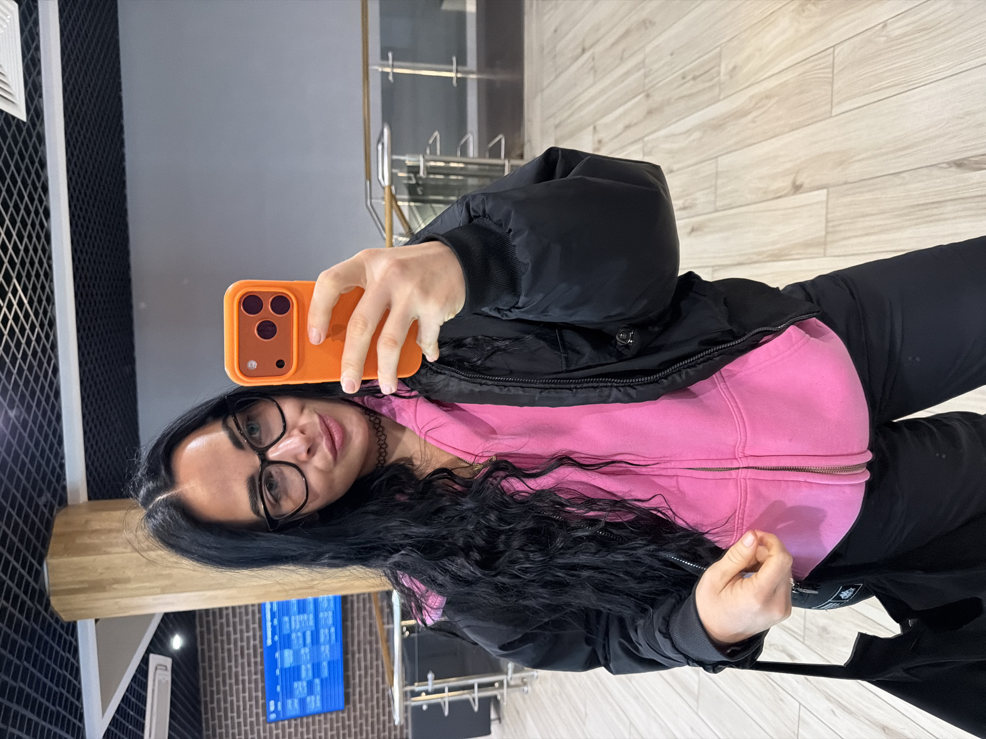 A woman is taking a selfie with an orange smartphone in a room that appears to be a public space. She is wearing glasses, a pink zip-up hoodie, and a black jacket. Her long, dark hair is styled in loose waves.
The background shows a modern interior with light-colored wood-look flooring, gray walls, and black patterned wall elements. There is also a visible metal railing system and a blue information board.
Without additional information, pinpointing the exact location within Saint Petersburg, Russia, is not possible.
