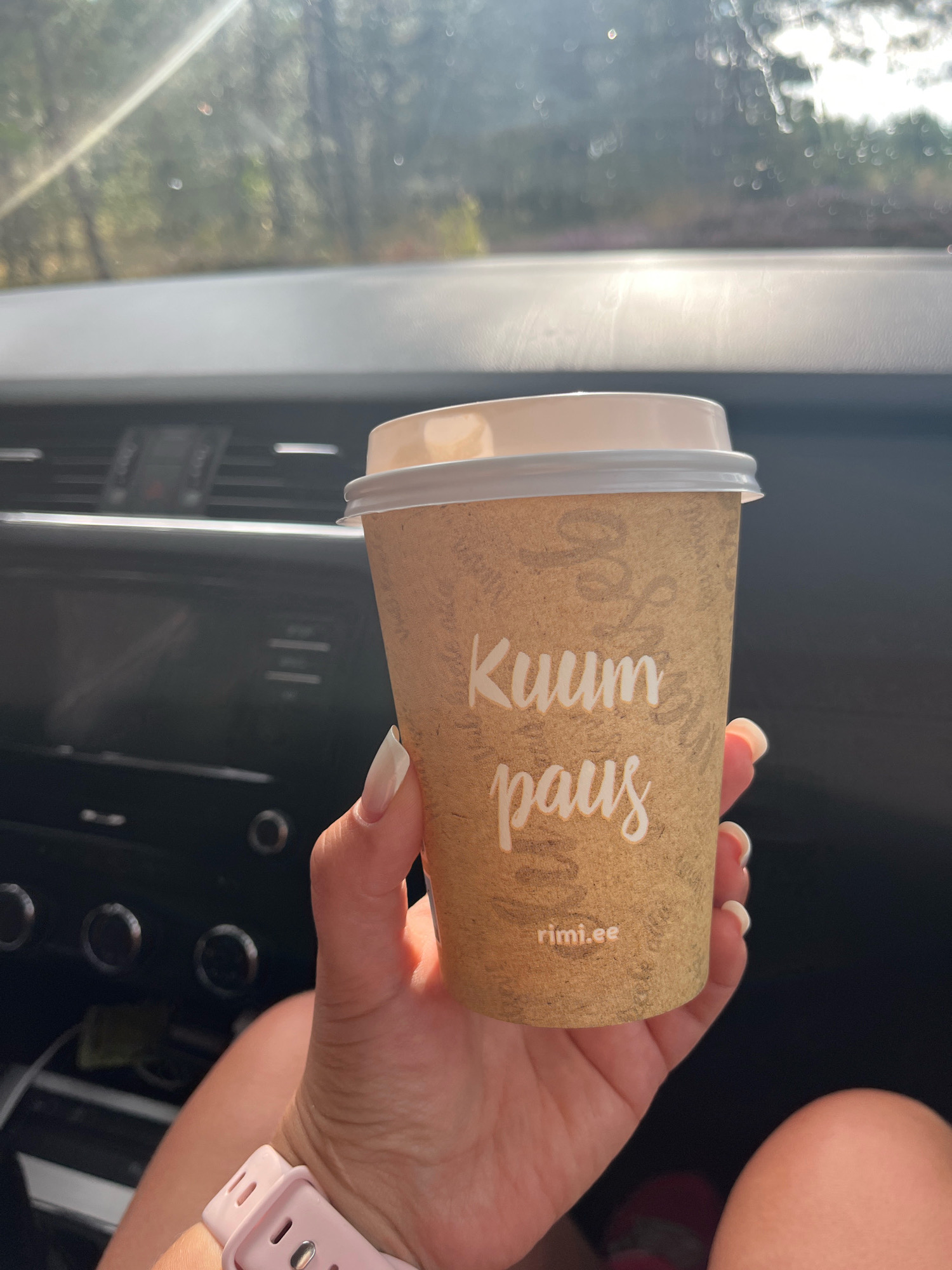 A person's hand holds a brown paper coffee cup with a white lid. The cup has "Kuum paus" and "rimi.ee" printed on it in white text. The background shows the interior of a car, with a dashboard and a sunlit view through the windshield of trees and sky. The person is wearing a light pink smartwatch on their wrist. The scene appears to be within the city of Kaberneeme, Estonia, given the presence of the rimi.ee domain name, which is a supermarket chain in Estonia.