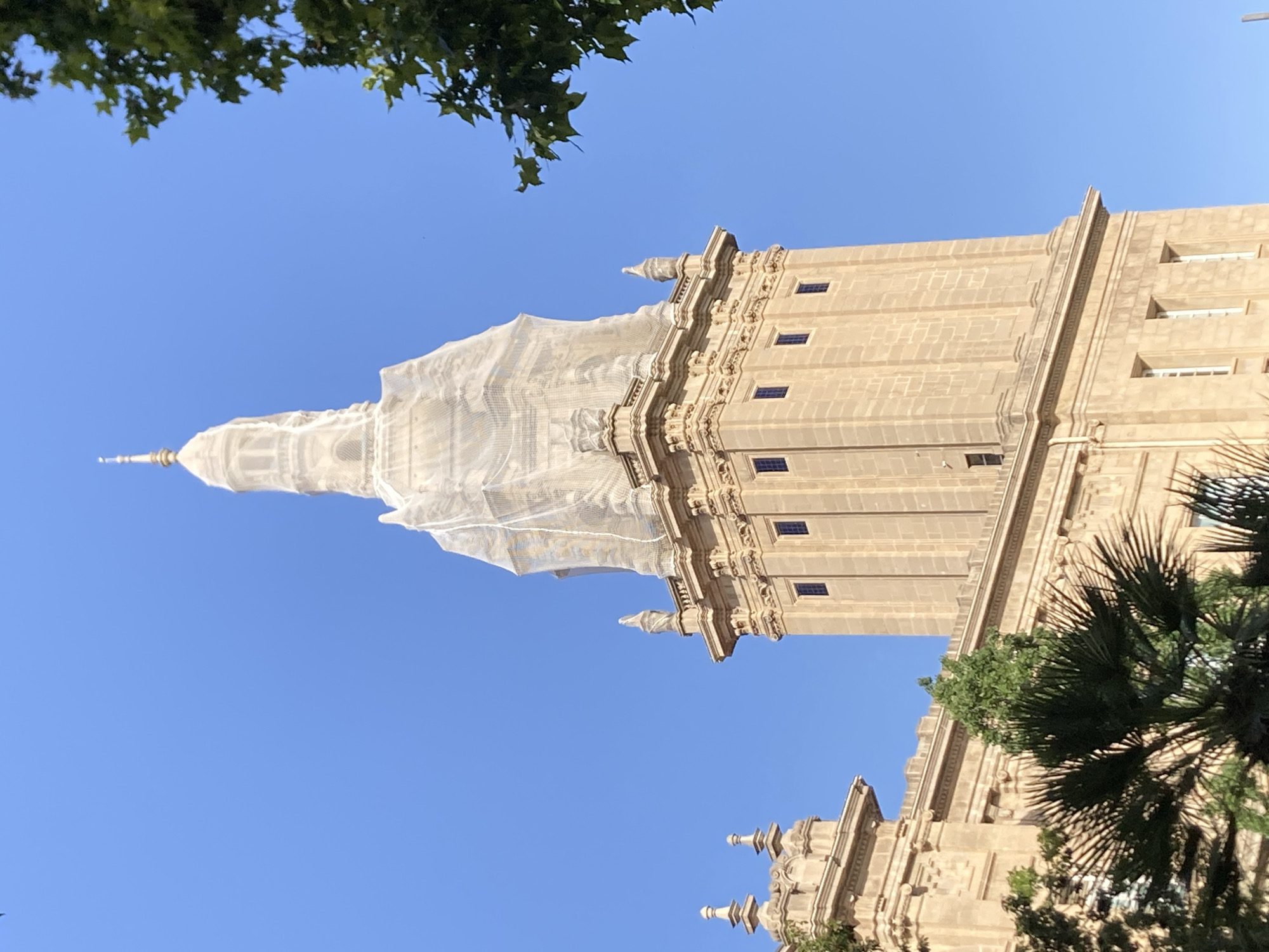 This image depicts the Palau Nacional, a grand palace located on Montjuïc hill in Barcelona, Spain. The palace, constructed between 1926 and 1929, is a prominent landmark and currently houses the Museu Nacional d'Art de Catalunya (MNAC). The photograph focuses on the upper portion of one of the palace's towers, which is partially covered in a white protective netting, suggesting ongoing restoration or maintenance work. The tower is made of light-colored stone and features several rectangular windows. Architectural details, such as ornate carvings and finials, are visible along the tower's facade. In the foreground, lush green foliage from trees and palm trees partially obscures the lower sections of the building. The sky is a clear, vibrant blue, indicating fair weather. No people are visible in the image.