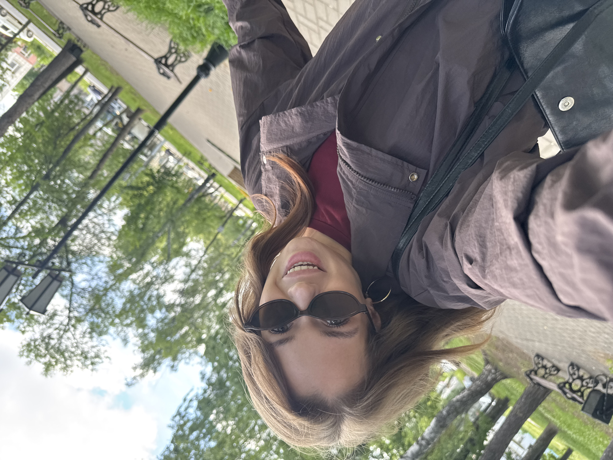 A young woman with light brown hair and sunglasses is smiling at the camera in a selfie. She is outdoors in a park with trees and benches. A street lamp is visible in the background. The image is taken from a low angle and appears to be in portrait orientation. The woman is wearing a brown jacket over a maroon shirt. She is carrying a black handbag. The park appears to be paved with cobblestones and lined with grass. The overall impression is that of a pleasant day in a public park. The location is identified as the city of Chaikovsky, Russia.