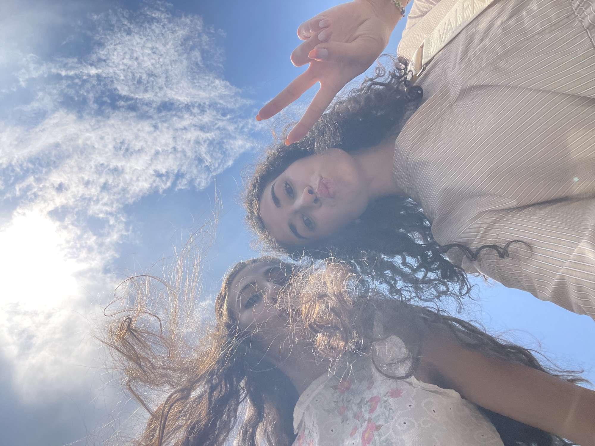 This is a first-person perspective, low-angle shot of two young women posing for a selfie against a bright blue sky with scattered white clouds. The woman on the right is in the foreground, making a peace sign with her right hand, her nails painted coral. She is puckering her lips and looking at the camera. She has dark, curly hair and is wearing a light brown, pinstriped top. The woman on the left is partially obscured by her own long, brown hair, which is being blown by the wind. She is wearing a white top with a floral pattern. The photograph is taken outdoors, suggesting a location with open sky. Given the information provided, it is not possible to determine the location within the city of Sofia, Bulgaria.