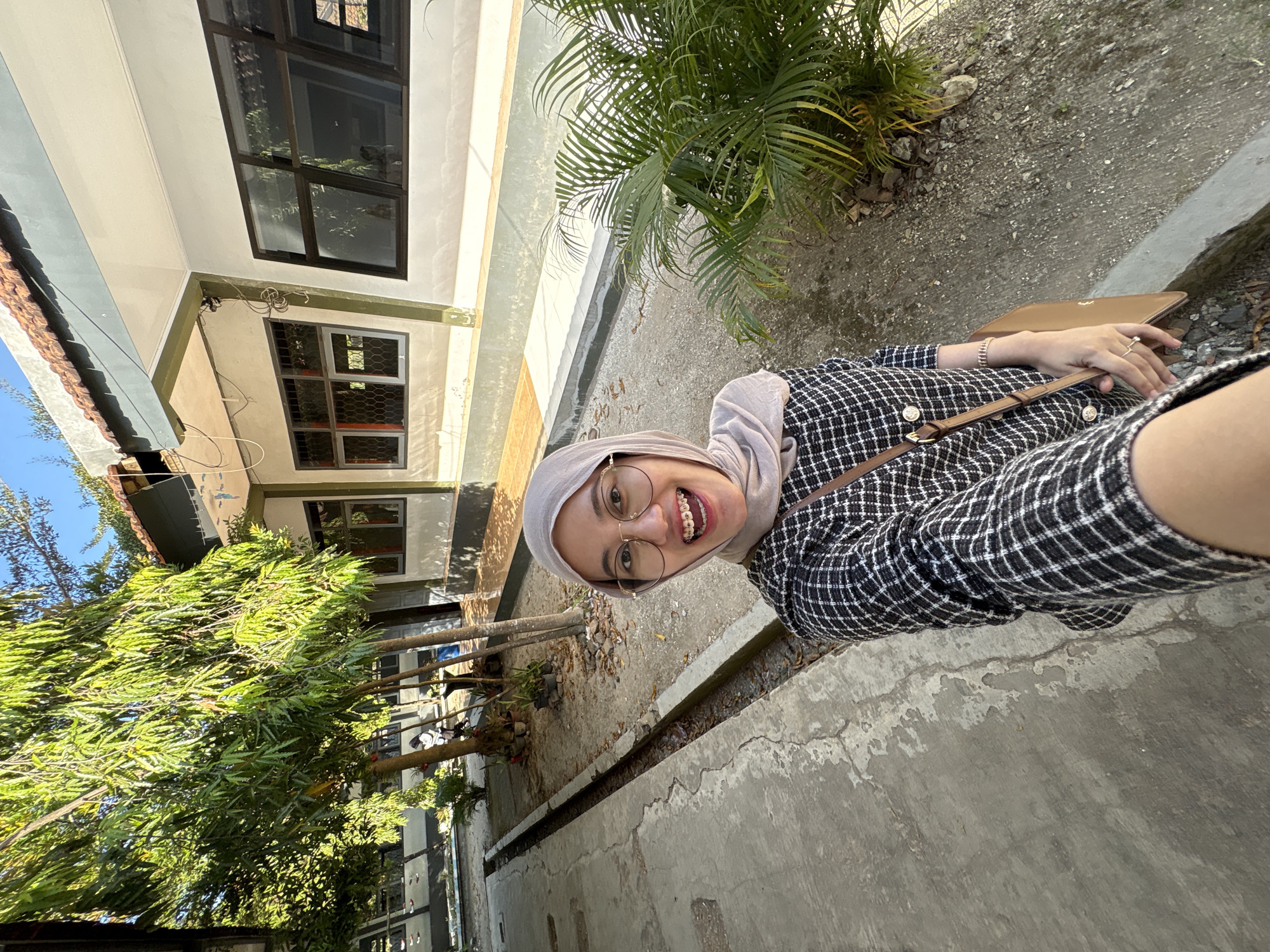 In Gunungsari, Indonesia, a young woman with a headscarf and glasses is taking a selfie outdoors on a sunny day. She is wearing a black and white checkered jacket and has braces on her teeth. She is holding a brown purse. The background shows a building with windows, trees, and a paved walkway.