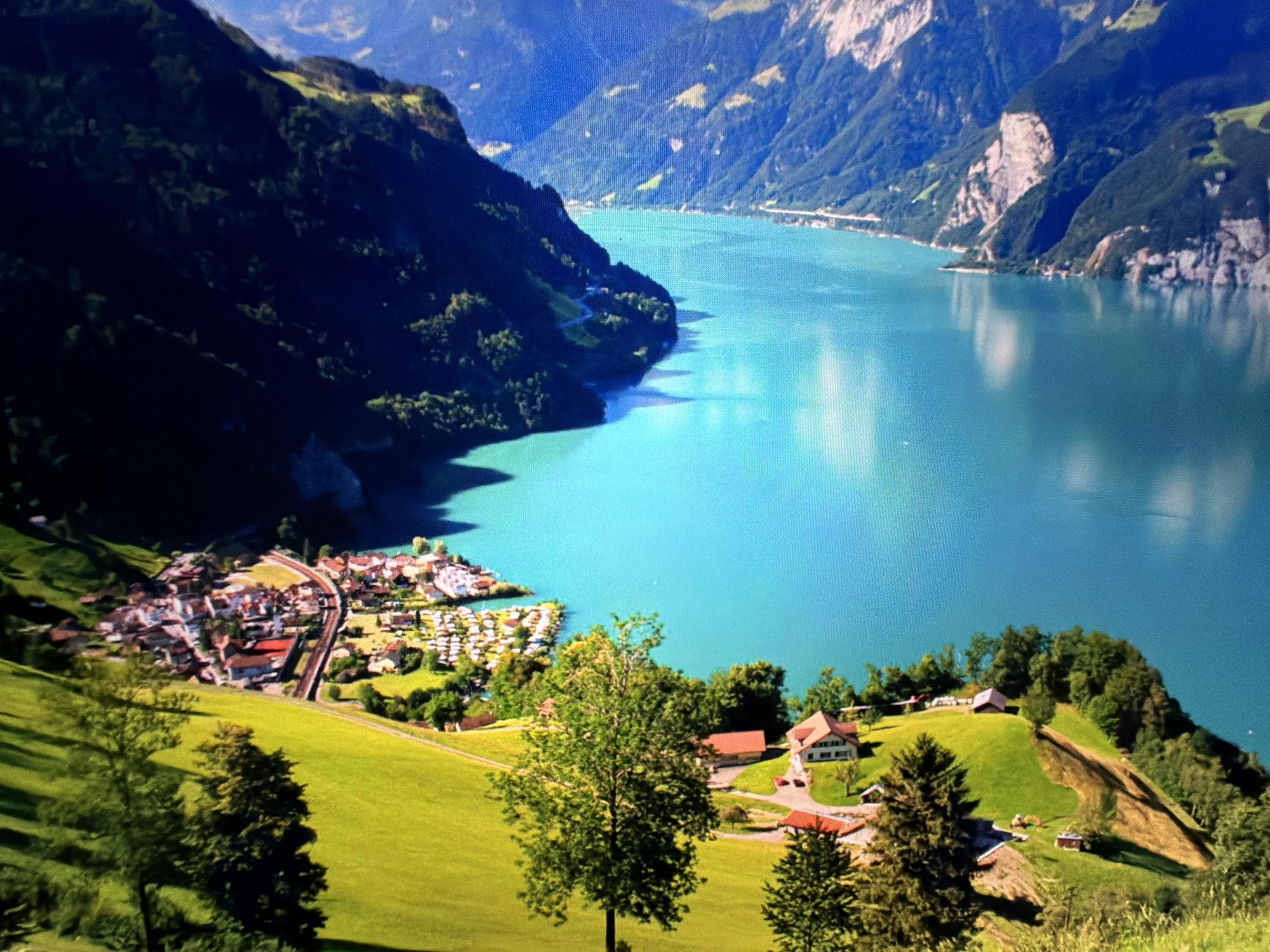 A high-angle, wide shot captures a vibrant, turquoise lake nestled among steep, dark green mountains. The lake's surface reflects the bright blue sky, dotted with white clouds. In the foreground, a lush, green meadow slopes downwards, featuring scattered trees and a winding path. On the left side of the meadow, a charming village with red-roofed houses and white buildings is situated along the lake's edge. A railway track curves through the village, and a campground with numerous white tents is visible nearby. Further along the lake, another cluster of houses and trees adorns a gentle incline. The scene is bathed in bright sunlight, casting shadows and highlighting the vivid colors of the landscape. There are no people visible in the image.