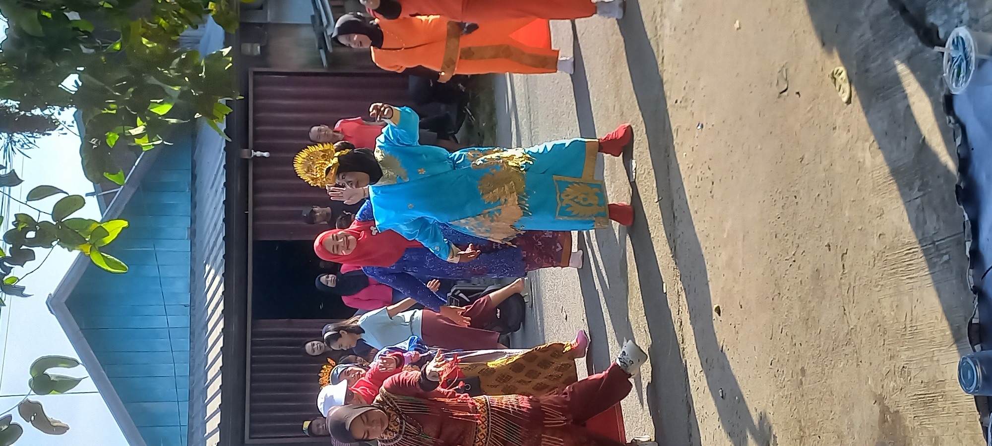 A group of people are gathered outdoors, possibly for a cultural event or celebration, in what appears to be a street in Samarinda, Indonesia. The central figure is a woman in a bright blue traditional costume adorned with gold detailing and a golden headdress, actively dancing with her arms outstretched. To her left, a woman in a red hijab and blue patterned dress is smiling and also appears to be dancing. To the right of the central dancer, another woman in an orange traditional outfit is visible. Other individuals, some in traditional attire and others in casual clothing, are observing the performance. The background shows a building with a corrugated metal facade and a light blue, angled roof, suggesting a simple urban or semi-urban setting. Trees with green leaves overhang the scene, casting shadows on the concrete ground, indicating it is daytime with bright sunlight. The presence of a few scattered items on the ground, like what might be discarded trash or debris, hints at an informal public space.