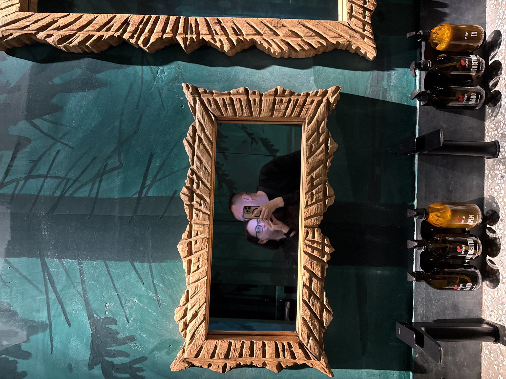 A couple is visible in a selfie reflected in a wooden-framed mirror. The background wall is painted in shades of teal with dark green leaf or plant-like patterns. Above the mirror, a portion of another similar wooden frame is visible. To the right, there are three dispenser bottles mounted on a black surface with a black faucet and what looks like a light-colored speckled countertop. Based on the lettering on the bottles, it is inferred that the location is Kazan, Russia.