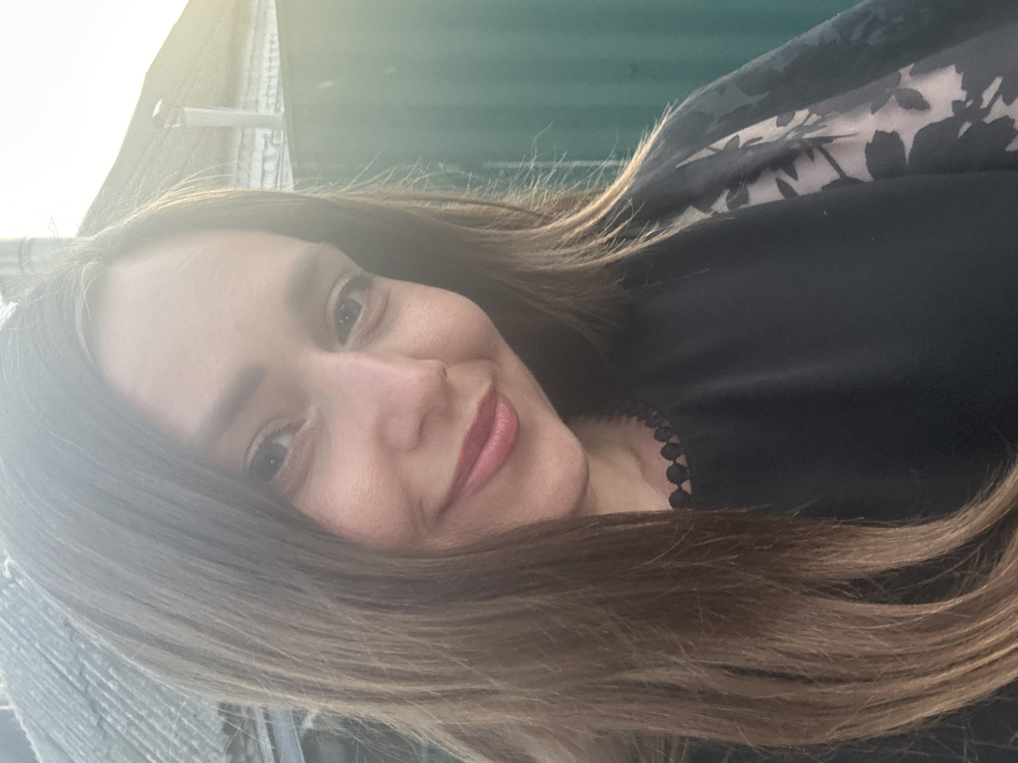 A close-up, outdoor selfie features a woman with long, brown hair and fair skin. She is smiling, and her lips are painted with a pink lipstick. She is wearing a black top with a lace trim around the neckline and sheer sleeves with a floral pattern. The background is blurred, but appears to be a residential area with a green fence, a roof, and a bright sky with sunlight casting a lens flare across the image. The location is not identifiable from the image itself.
