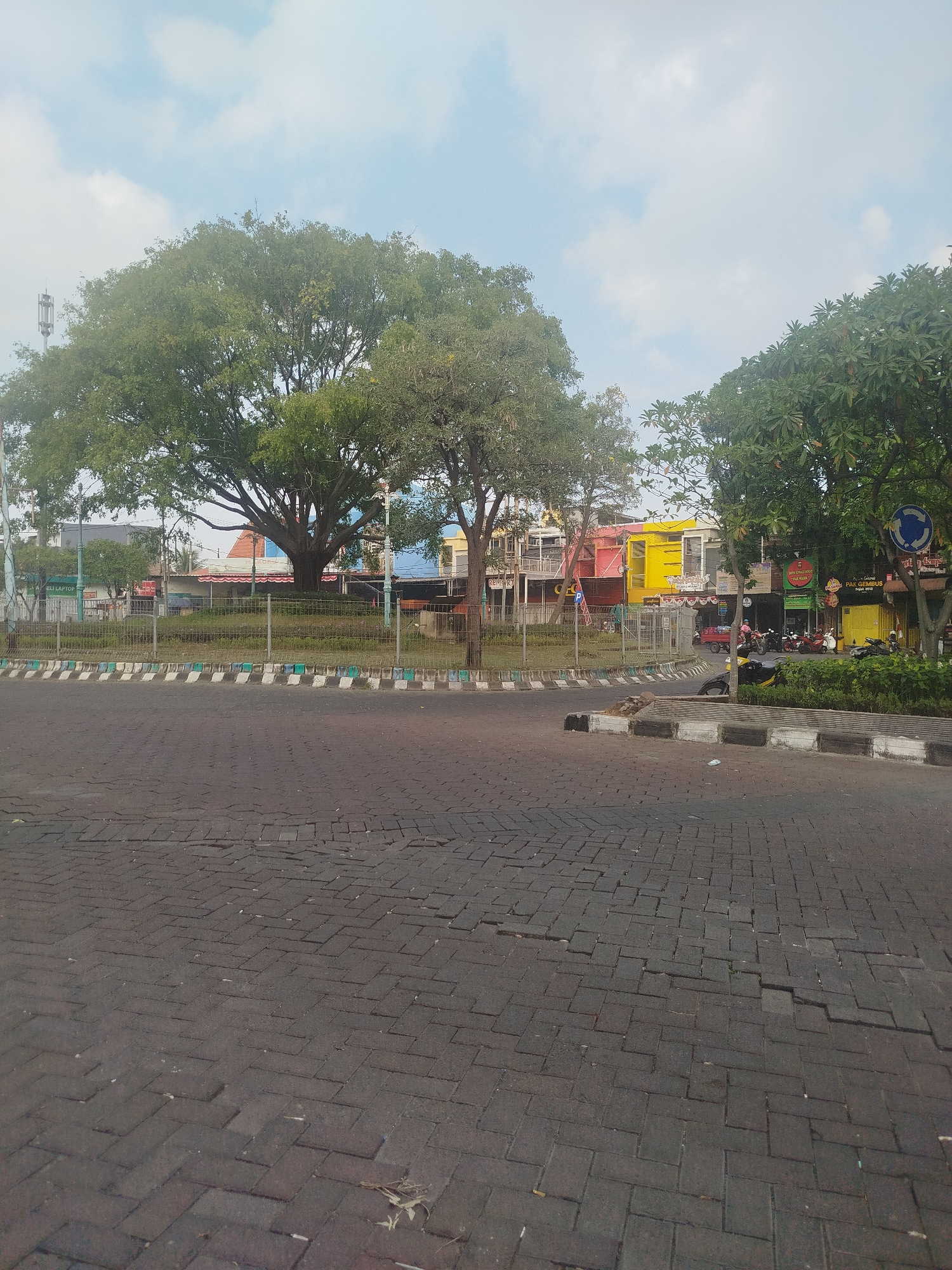 The image depicts a street intersection in Yosowilangun, Indonesia. The foreground is dominated by a paved road made of dark grey interlocking bricks, showing signs of wear and some cracking. A low, decorative curb with alternating white and green/blue bands runs along the edge of the road. Beyond the curb is a grassy median surrounded by a metal fence.
In the middle ground, a large, mature tree with a broad canopy of green leaves stands prominently. Behind the tree, several buildings are visible, exhibiting a range of colors, including yellow, pink, and blue. Some of these appear to be shops or businesses, with signage visible. Several motorcycles are parked in front of the buildings. A red utility vehicle is also present on the street.
The background features more trees and a clear sky with scattered white clouds. A cell tower is visible to the left of the large tree. The overall scene suggests a somewhat urban environment, likely a commercial area within the town. There are no people clearly depicted in the image, although the presence of parked vehicles implies recent activity. The lighting suggests it is daytime, possibly morning or late afternoon, given the soft light.