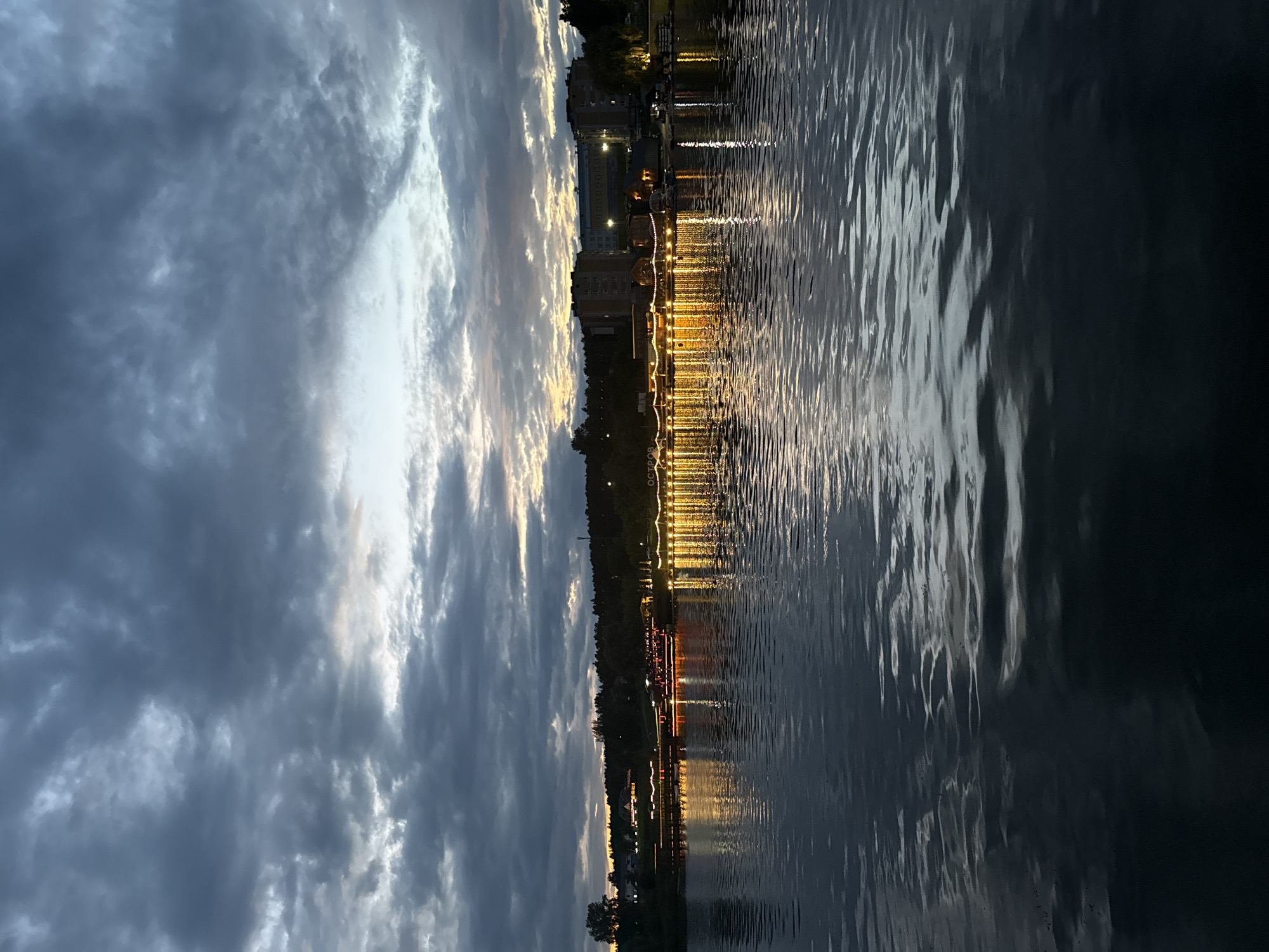 The image depicts a twilight scene over a body of water, likely the Votkinsk Reservoir in Votkinsk, Russia. In the distance, a shoreline is lined with trees and several buildings, including apartment blocks. A string of lights illuminates a long pier or embankment extending along the water's edge, reflecting brightly on the rippling surface of the water. The sky is filled with dramatic, dark clouds, with a sliver of lighter sky visible near the horizon, suggesting the last vestiges of daylight. No people are visible in the image.