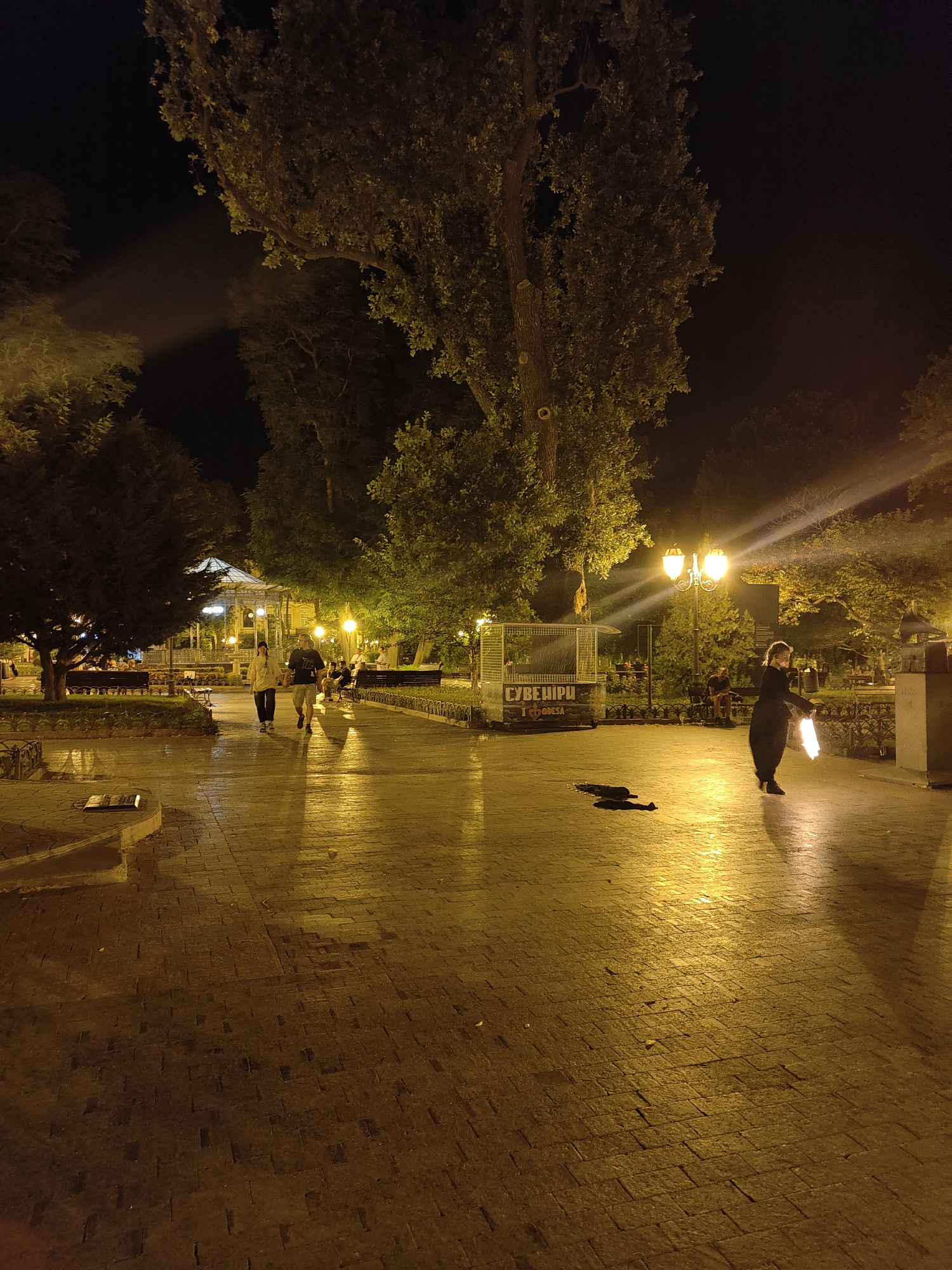 In the city of Odesa, Ukraine, a person is performing with fire at night in an illuminated park. The ground is paved with dark bricks, reflecting the warm light from street lamps and a large tree. Several people are walking in the distance, some sitting on benches. A woman in a dark outfit is holding two flaming objects, with flames emanating from them. The background shows trees and a building with a glass roof. The scene is lit by multiple street lamps, creating long shadows and highlights on the wet pavement. A small cage-like structure with text on it is visible to the left of the performer.