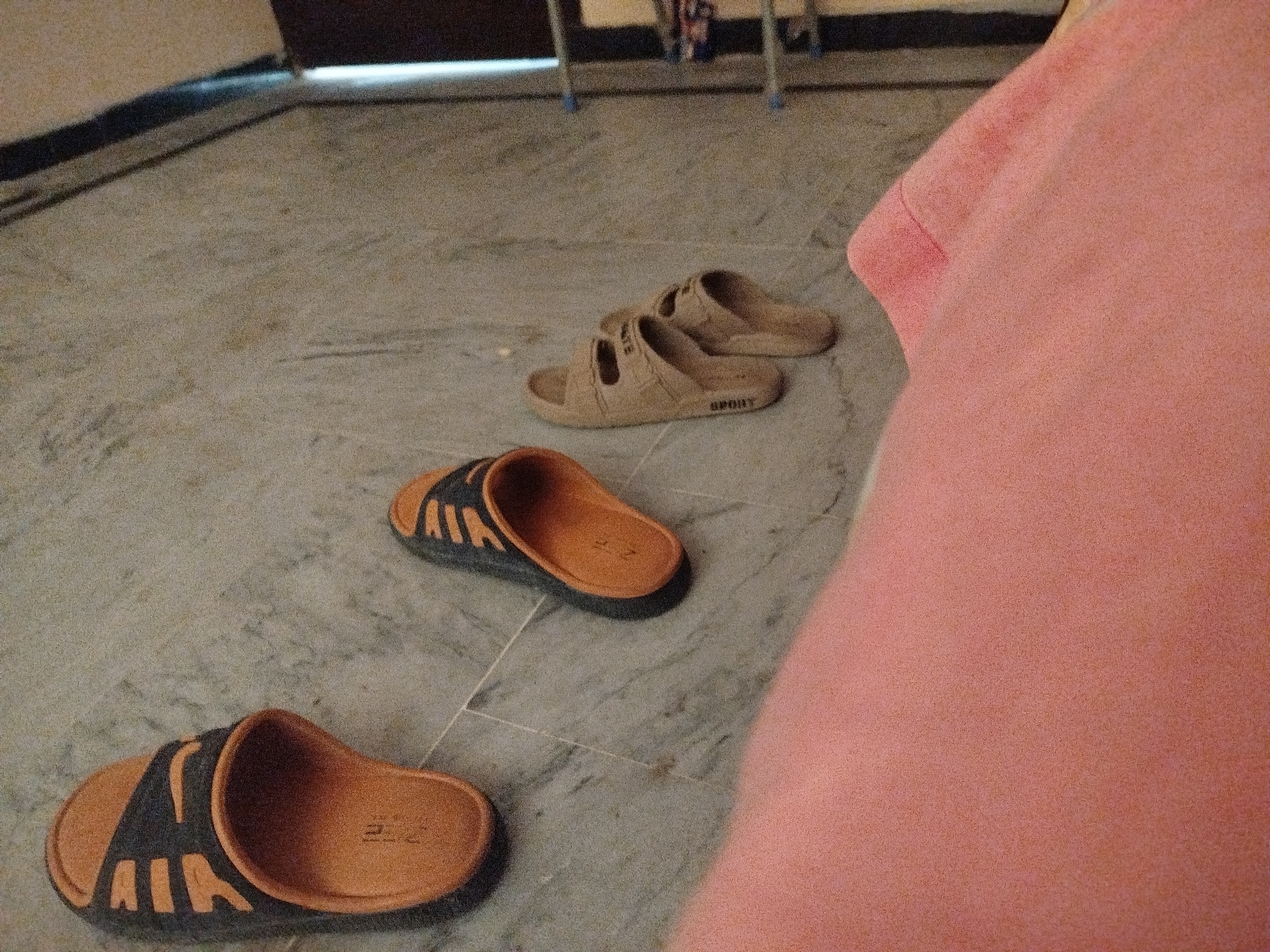 The image shows a close-up, overhead view of three pairs of slippers on a marble-tiled floor. Two pairs are tan and one pair is orange and black. The slippers are arranged in a diagonal line from the bottom left to the middle right of the frame. A pink garment, possibly clothing, is prominently displayed on the right side of the image, obscuring a portion of the floor and the slippers. In the background, a metal stand with blue plastic parts is visible. The scene appears to be indoors, in what could be a residential setting. No specific location in the city of Lassan, Pakistan is identifiable from the visual information provided.