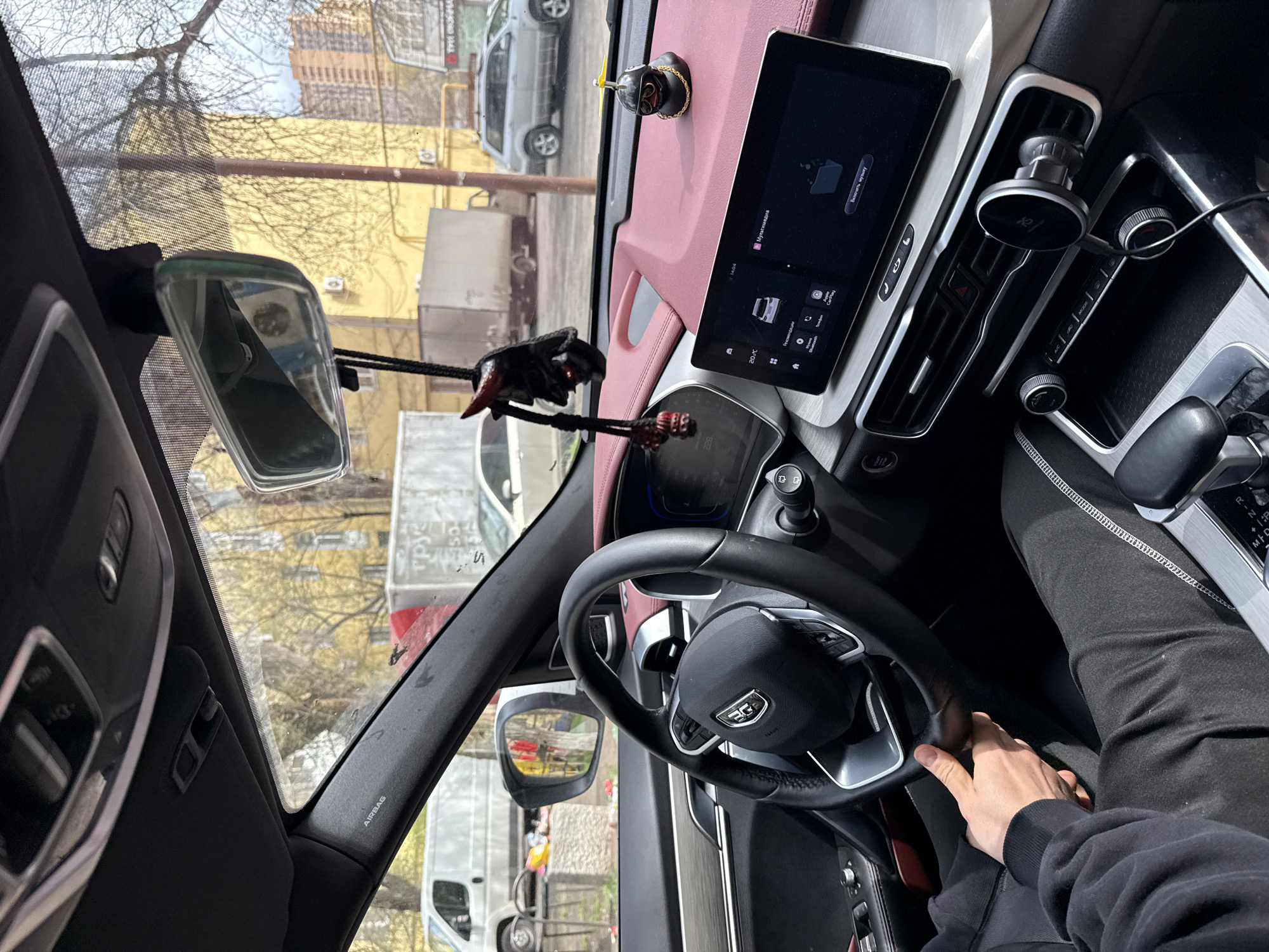 The image depicts the interior of a car, likely in Rostov-on-Don, Russia, based on visible signage in the reflection of the mirrors. The dashboard is a combination of pink leather and gray. The steering wheel is black and bears a logo, held by a right hand. The center console features a display screen showing what appears to be car settings in Russian. Through the windshield, a street scene is visible, with buildings and vehicles, including delivery vans bearing Russian text. A rear-view mirror and other dashboard ornaments are visible.
