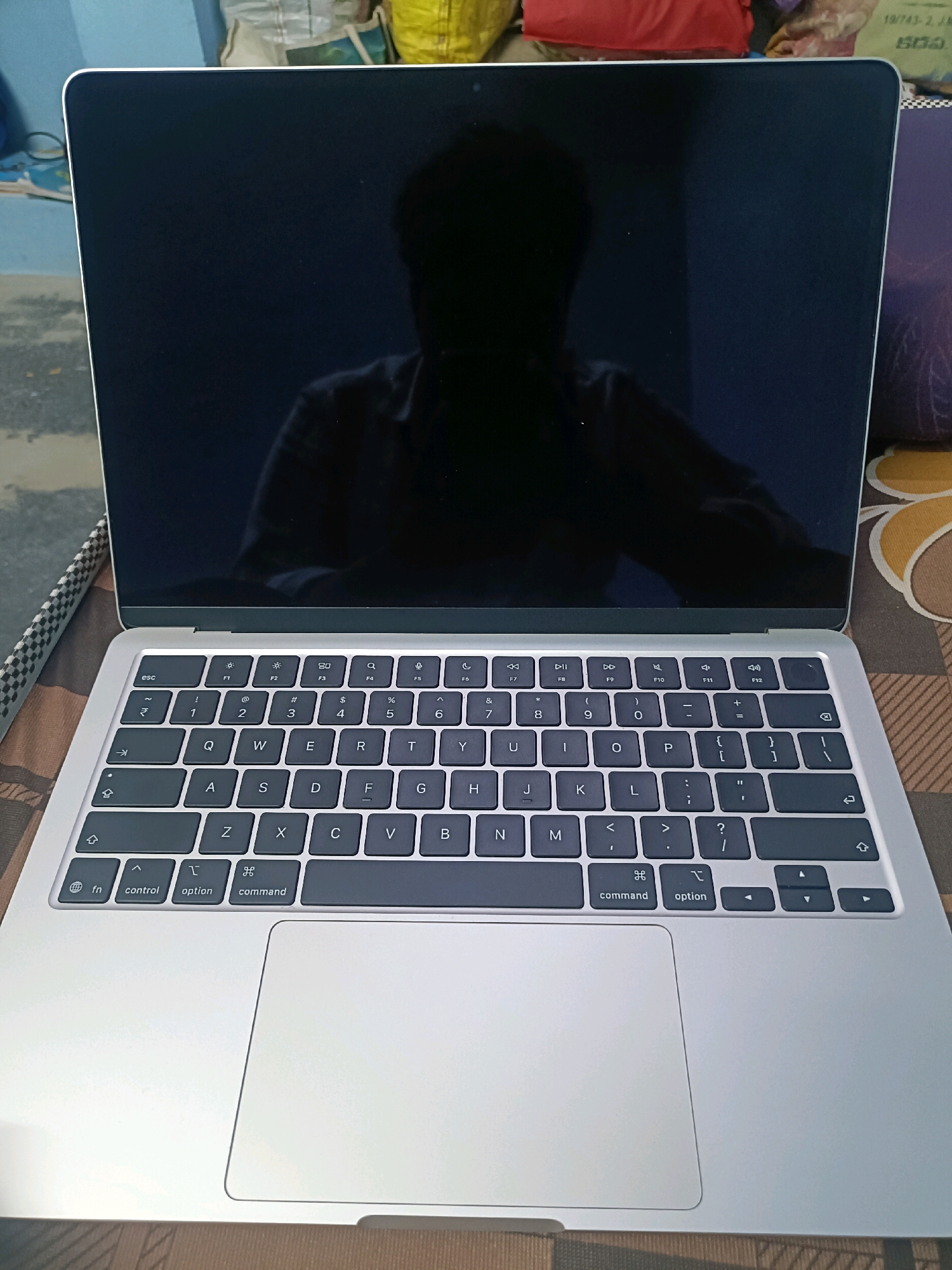 The image shows a silver MacBook laptop with its screen open. The screen displays a dark reflection, possibly of the person taking the photo. The keyboard is visible below the screen, showing standard QWERTY layout along with function keys and special keys like 'fn', 'control', 'option', and 'command'. The laptop is placed on a patterned surface, which appears to be a mat or blanket. In the background, there are out-of-focus objects, including what look like some bags and a blue structure. There is no information in the image to indicate the location in the city of Puttampalli, India.