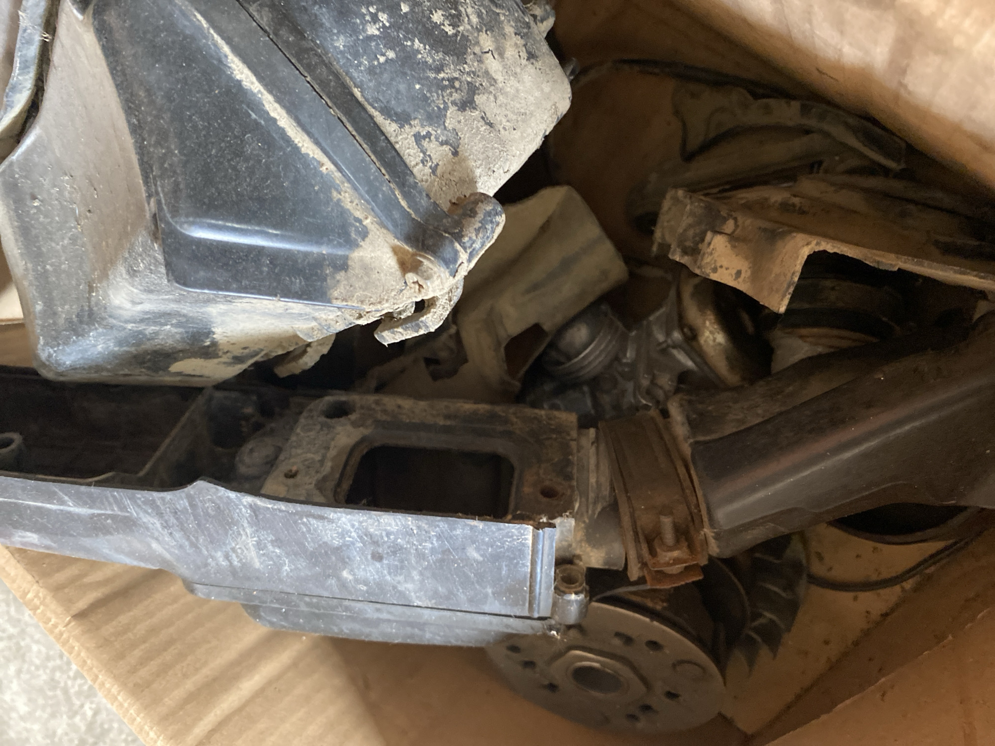 The image shows a close-up, overhead view of a cardboard box filled with various dusty, dirty, and possibly used automotive or mechanical parts. The items are jumbled together, with no clear arrangement. Visible components include what appear to be plastic housings, metal brackets, a circular metal disc with holes, and other unidentifiable pieces of machinery. The lighting is natural and appears to be coming from above, casting shadows within the box. The environment is utilitarian and appears to be an interior space, likely a workshop or storage area. There are no people visible in the image. The origin of the image, "city of Bayu, Indonesia," is not visually ascertainable from the provided photograph.