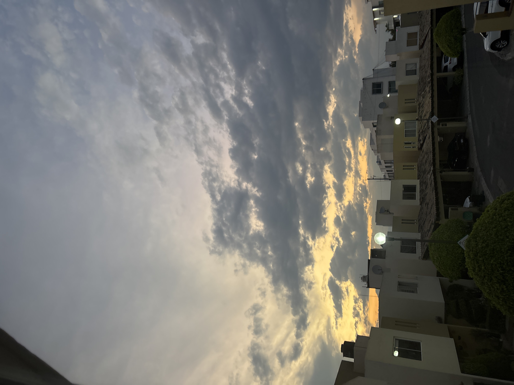 The image captures a view of the sky and adjacent buildings, likely taken in Puebla, Mexico. The sky features a mix of light and dark clouds, with some areas illuminated by what appears to be the setting sun. The buildings are a light color, likely white or light beige, and appear to be part of a residential area. Street lights are visible near the buildings. Cars parked on the road suggest that this could be a local street in the city.