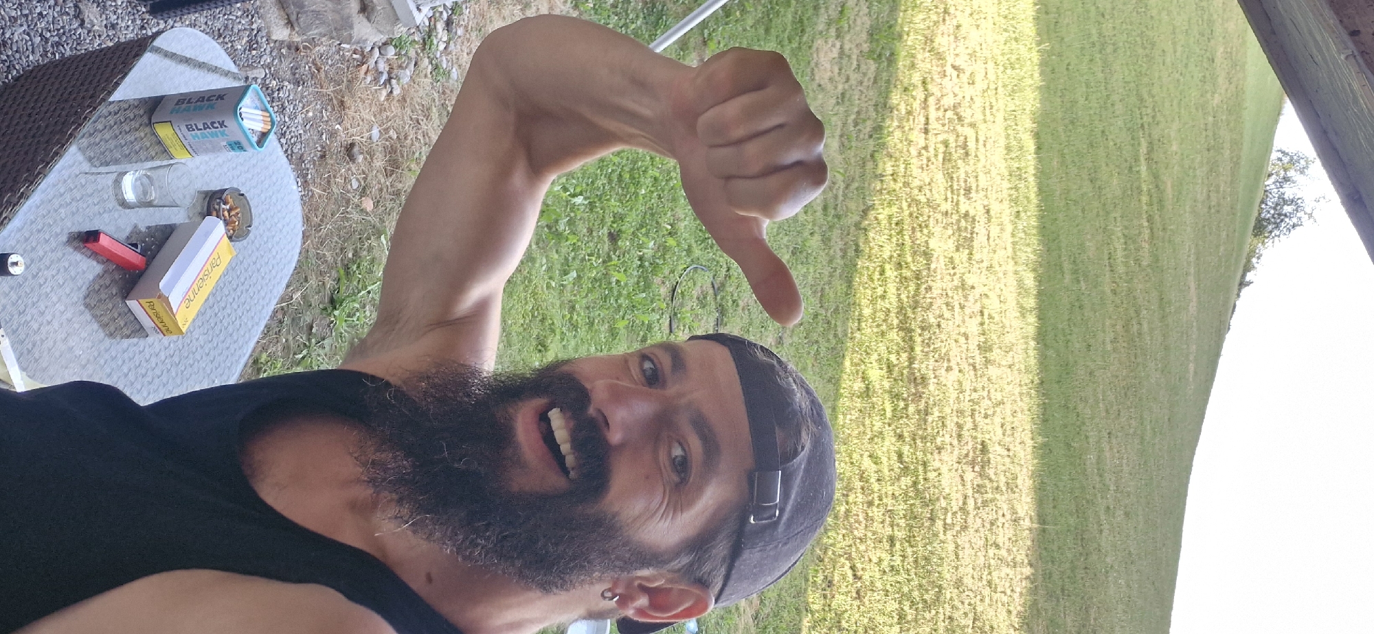 A man with a beard and a black baseball cap is posing for a selfie outdoors. He is wearing a black tank top and giving a thumbs-up gesture with his left hand. His right arm is flexed and visible. In front of him is a wicker table with a glass, an ashtray filled with cigarette butts, a cigarette pack labeled "Parisienne", and a red lighter. The background features a grassy hill and some trees. The man appears to be smiling.