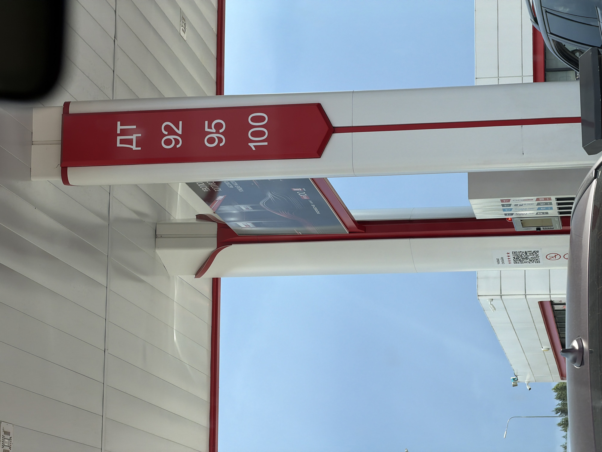 The image shows a gas station with a bright blue sky in the background. In the foreground, a white gas pump pillar is visible, featuring a red vertical stripe. On the pillar, the fuel types and prices are displayed in white text on a red background. The visible fuel types are "ДТ" (likely diesel fuel), "92", "95", and "100", with their corresponding prices. To the left of the pillar, a large advertisement is mounted, displaying an image of a car and text. Further to the left, the corner of a white building with a red stripe is visible, and to the right, part of a black SUV is in the frame. The overall scene suggests a typical gas station environment under natural daylight. The location is identified as Zhilotkovo, Russia, though this information is not visually verifiable from the image itself.