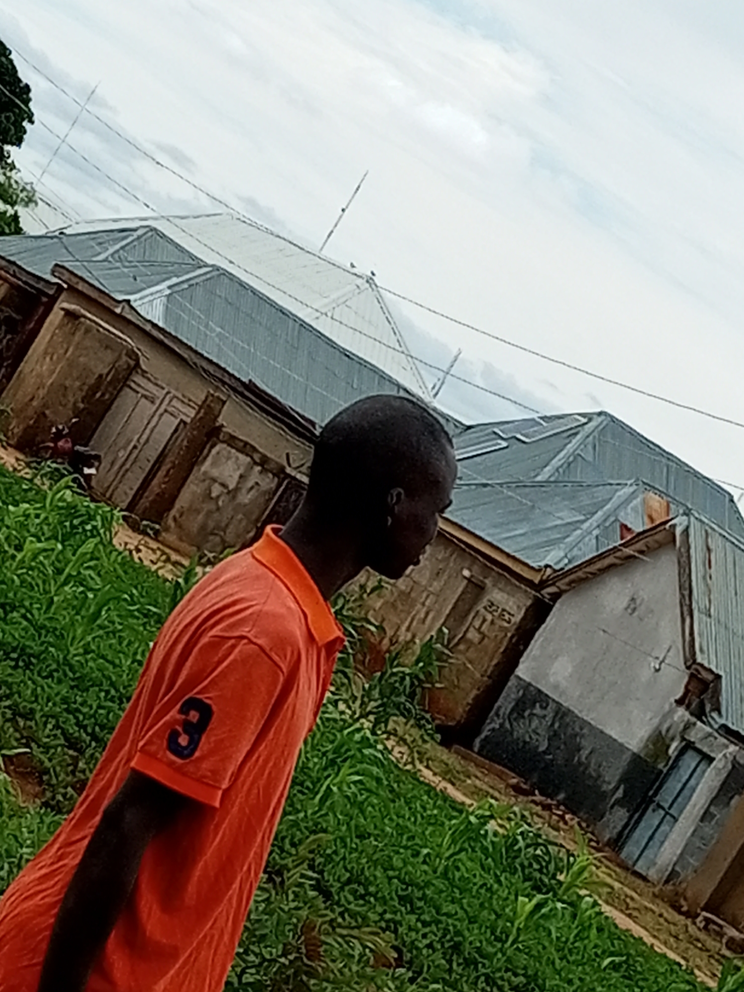In Tempure, Nigeria, a young Black male wearing an orange polo shirt with the number "3" on the sleeve stands in front of a row of buildings with corrugated metal roofs. The buildings appear to be constructed from various materials, including brick, concrete, and possibly wood. The overall scene depicts a somewhat disorganized and weathered urban environment. The sky is overcast, suggesting it may be a cloudy day. Lush green vegetation, possibly crops or weeds, surrounds the buildings and partially obscures the lower portion of the image. The perspective is slightly tilted, adding to the dynamic feel of the photograph.