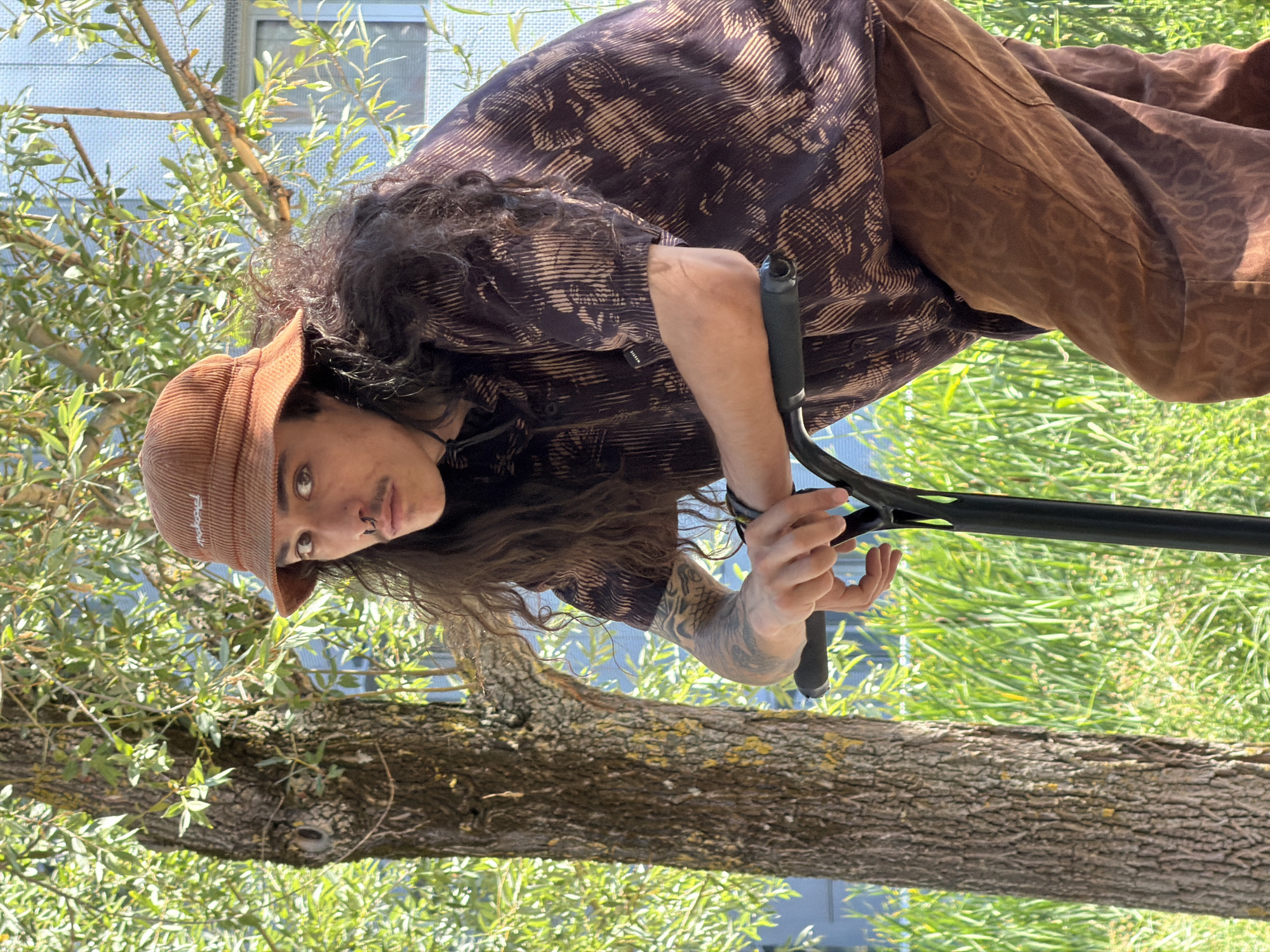 A person with long, dark, curly hair, a mustache, and a septum ring stands next to a tree, holding onto the handlebars of a black scooter. They are wearing a brown corduroy bucket hat, a dark shirt with a brown abstract pattern, and brown pants with a similar pattern. The background shows green grass and foliage, with a building visible in the distance. The lighting suggests it is daytime. The image appears to be taken outdoors, possibly in a park or urban green space. There is no identifiable indication of the location being Lyon, France.