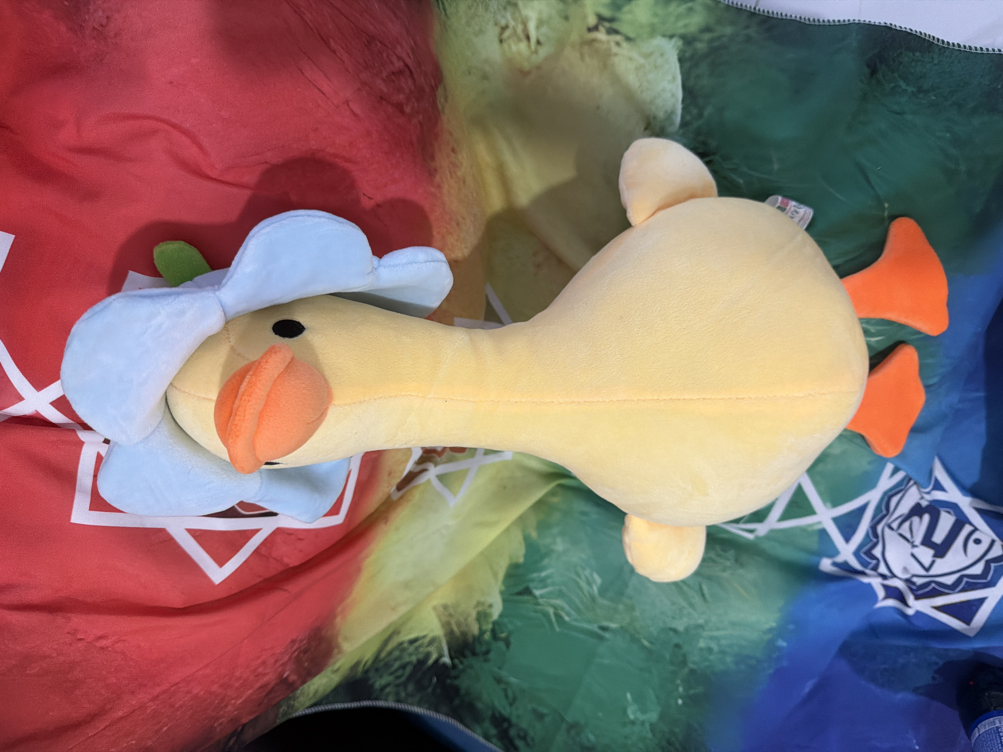 The image shows a yellow plush duck toy lying on a colorful, patterned fabric. The duck has a blue flower-like hat with a green stem on its head, and an orange beak and feet. The fabric beneath the duck appears to be a blanket or tablecloth with a gradient of colors from red on the left, through yellow and green in the middle, to blue on the right. There are white geometric patterns on the fabric, particularly visible on the red and blue sections. The lighting suggests it is daytime. The duck is the sole subject in the image, and no activity or event is taking place beyond the object being displayed. There is some faint, unreadable text on a white tag attached to the duck's body. The location context of Nha Trang, Vietnam, does not provide any specific visual cues within the image itself.