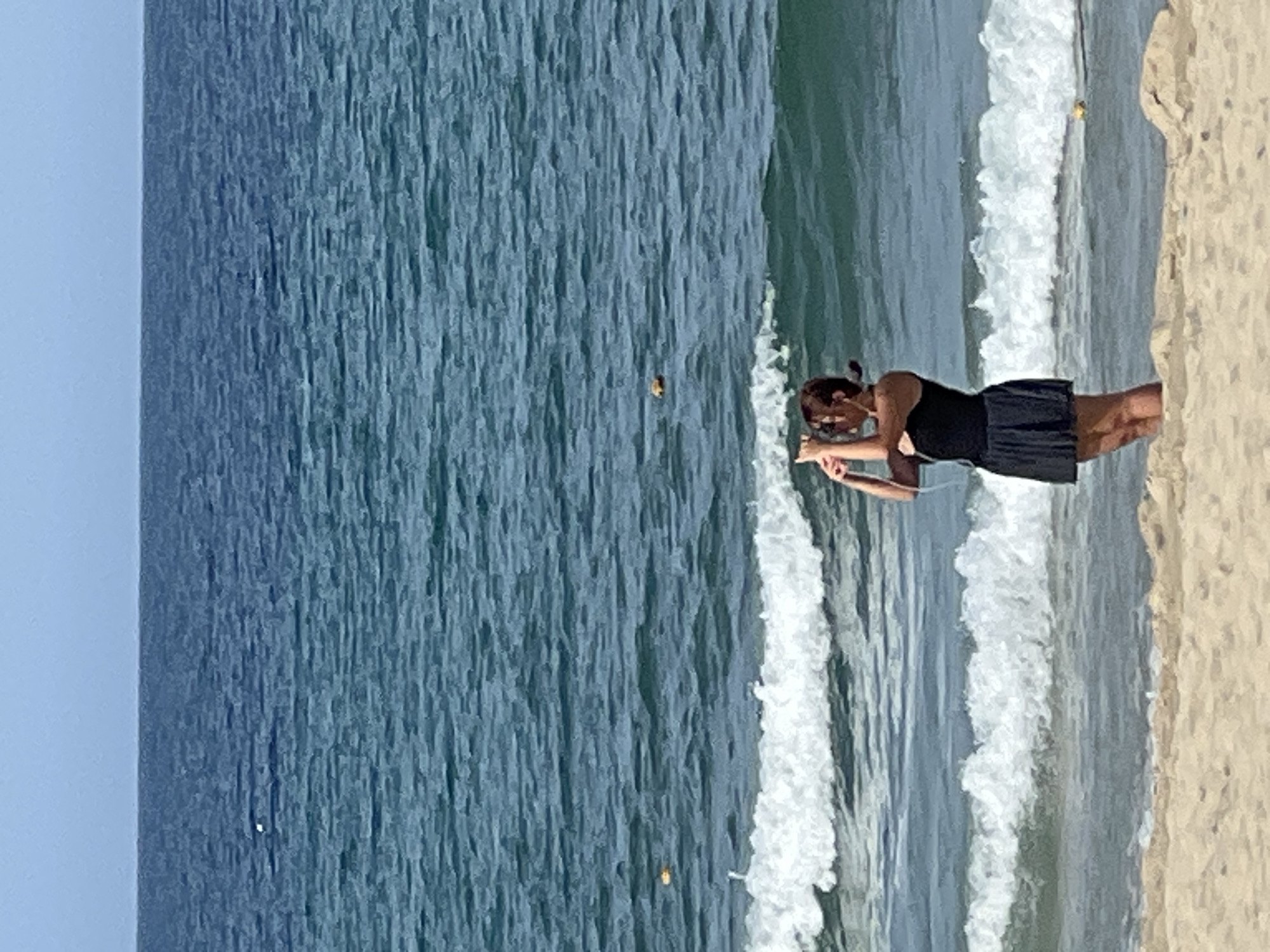 A young woman stands on a sandy beach at the coast of Đà Nẵng, Vietnam. She is wearing a black tank top and a black skirt. She is holding a smartphone in her hands and appears to be taking a picture or recording a video of the ocean. The ocean water is a deep blue, with white waves crashing onto the shore. The sky above is clear and pale blue. The overall scene depicts a casual moment of leisure at the beach.