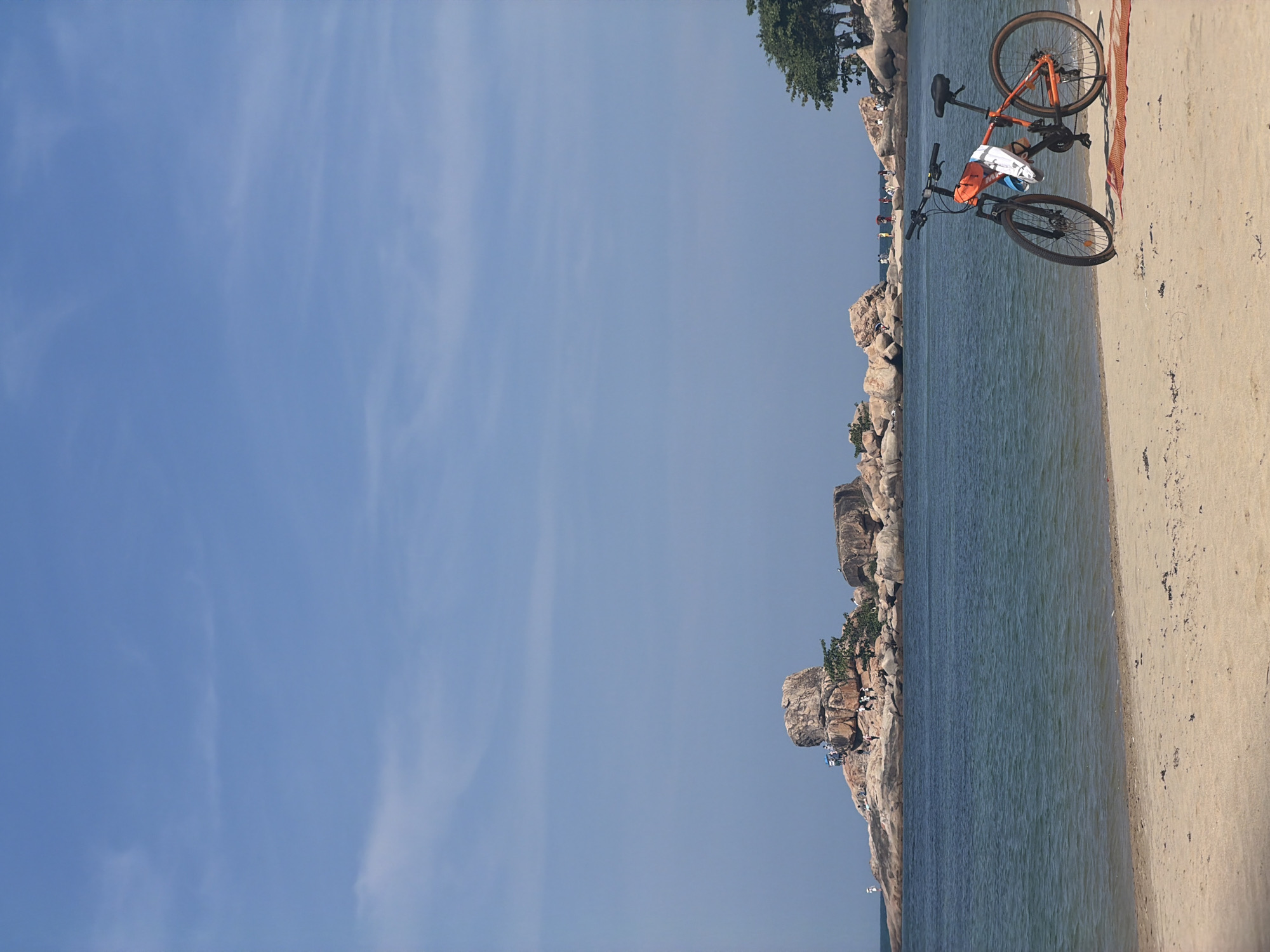 The image shows a shoreline scene in Bac Nha Trang Ward, Vietnam. An orange bicycle lies on a concrete surface next to a beach, with the sea stretching towards a rocky coastline. People are situated on the rocks. The sky is blue with scattered clouds.
