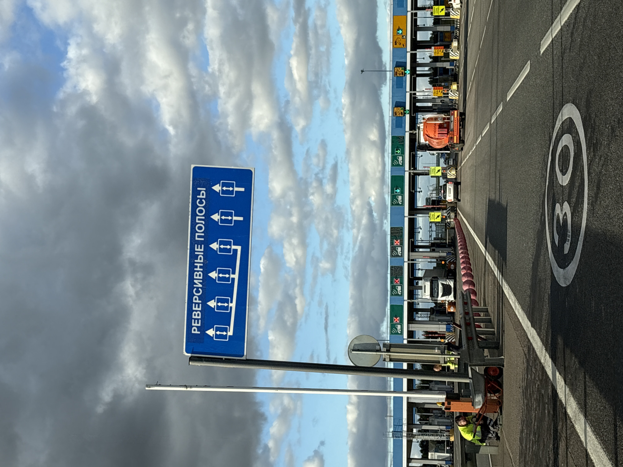 A road sign with blue background and white text reads "РЕВЕРСИВНЫЕ ПОЛОСЫ" which translates to "REVERSIBLE LANES". Below this text are five boxes, each containing two arrows pointing in opposite directions, indicating variable lane direction. The sign is mounted on a tall pole. In the foreground, a person wearing a high-visibility yellow jacket is crouched down next to a wheelbarrow. Another person in a similar jacket is standing nearby. They are positioned near a road barrier. The road ahead is a multi-lane highway leading to a toll plaza. The toll plaza has multiple booths, some of which are displaying green lights, indicating open lanes. Several vehicles, including a white truck and cars, are visible at the toll plaza. The road surface has white lane markings and a painted circle with the number "30" inside, indicating a speed limit. The environment appears to be outdoors with a cloudy sky. The scene is located at the toll plaza on the M11 Neva highway, near Ploty, Russia.
